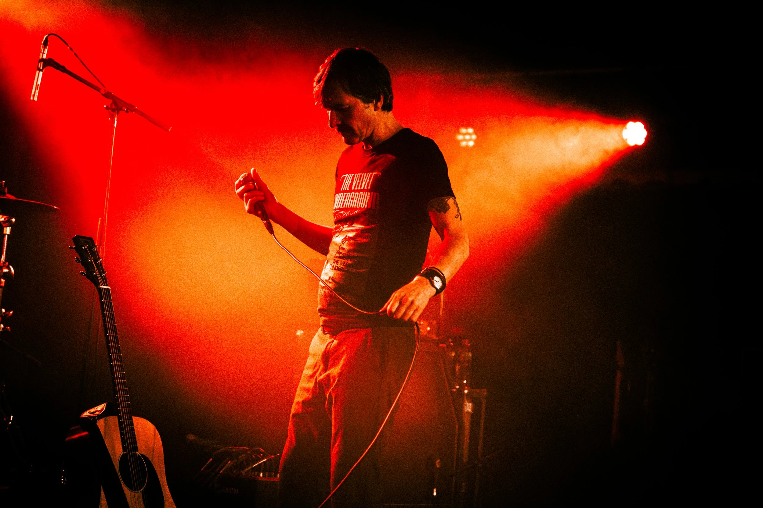 A male musician holding a microphone on stage, illuminated by red stage lighting, with a guitar placed on a stand nearby.