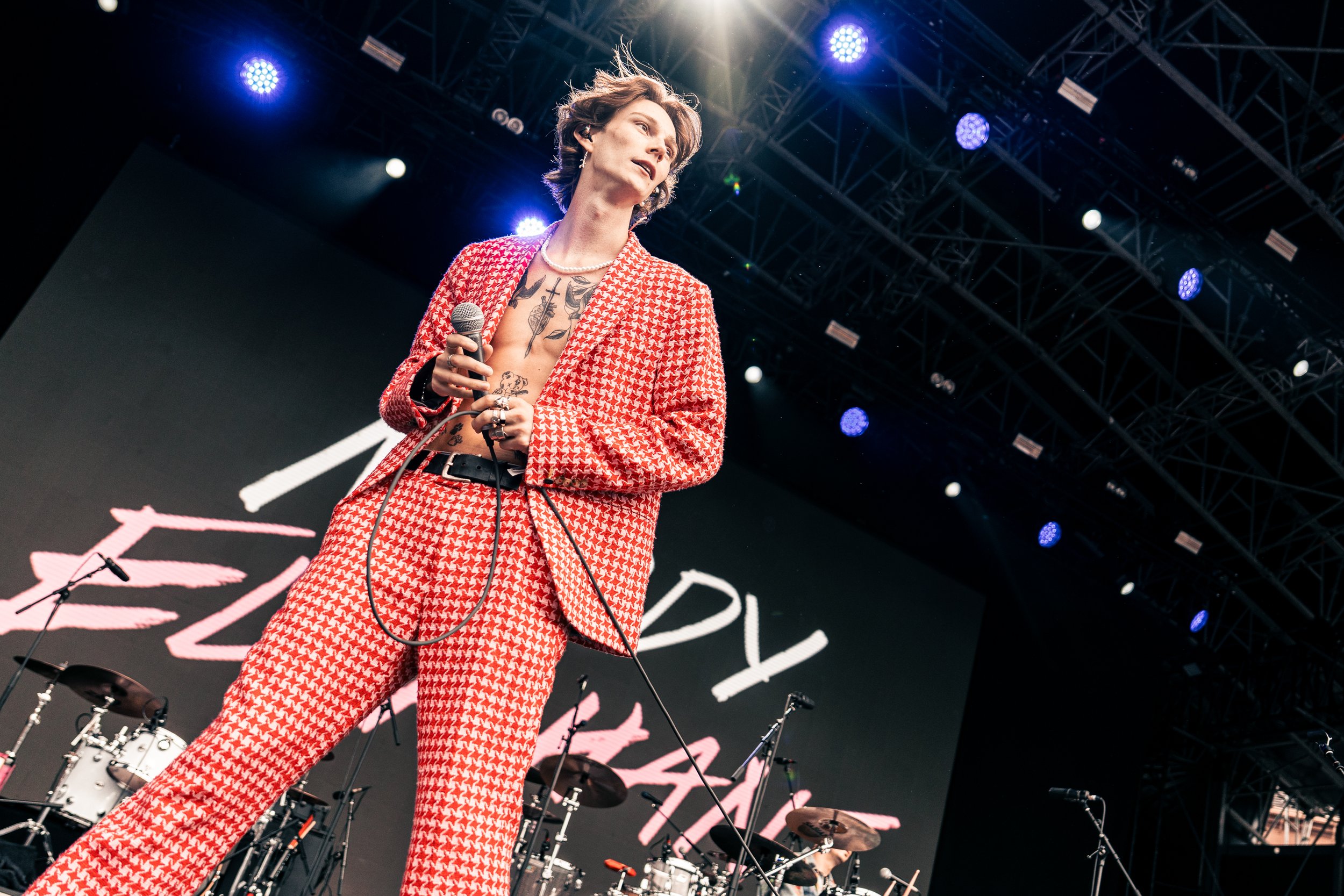 A woman on stage holding a microphone wearing a red and white patterned suit with tattoos visible on her chest, performing under stage lights with a large screen behind her and musical instruments around.