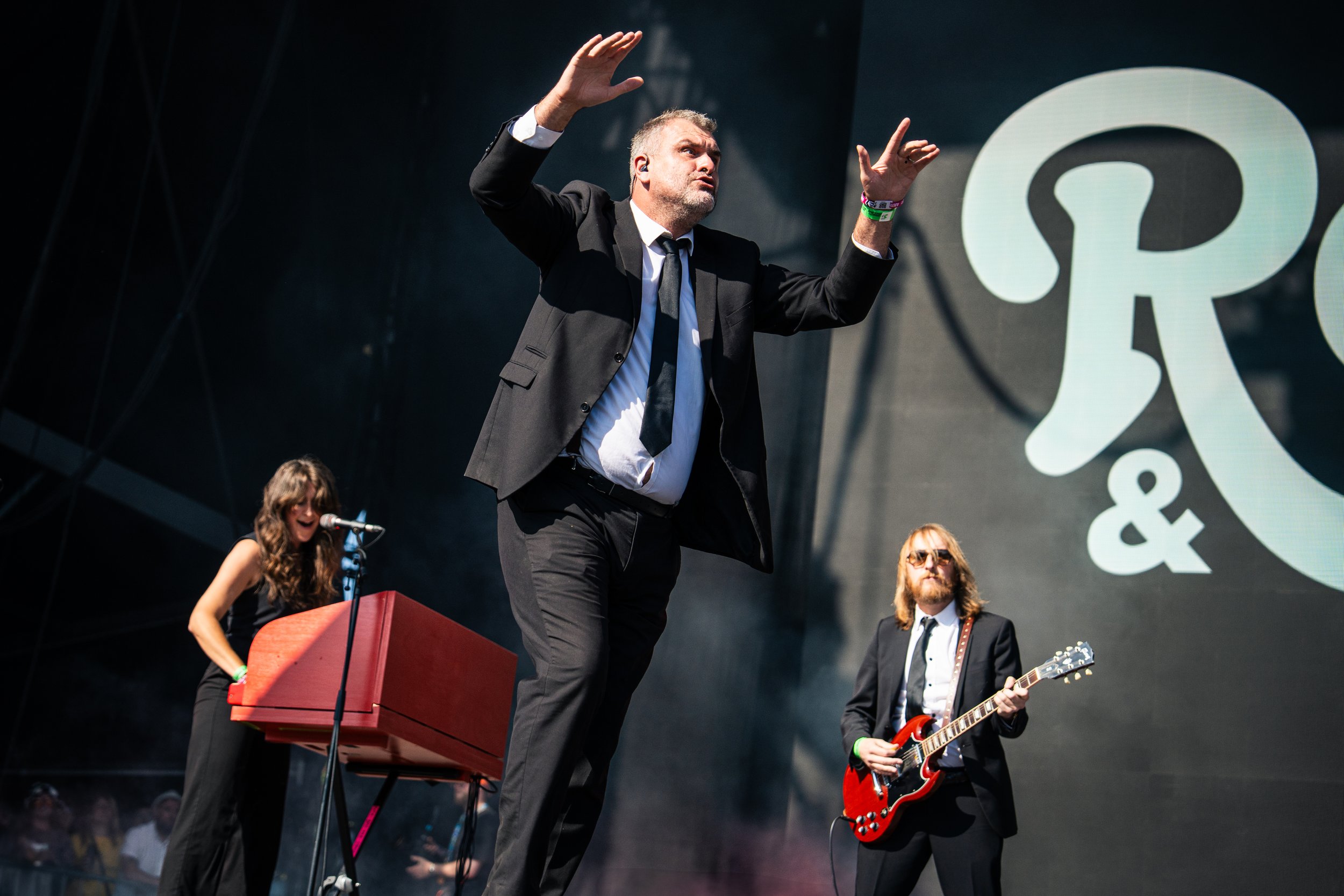 Band performing on stage, man in a suit singing, woman playing keyboard, man with guitar, large screen behind with logo.