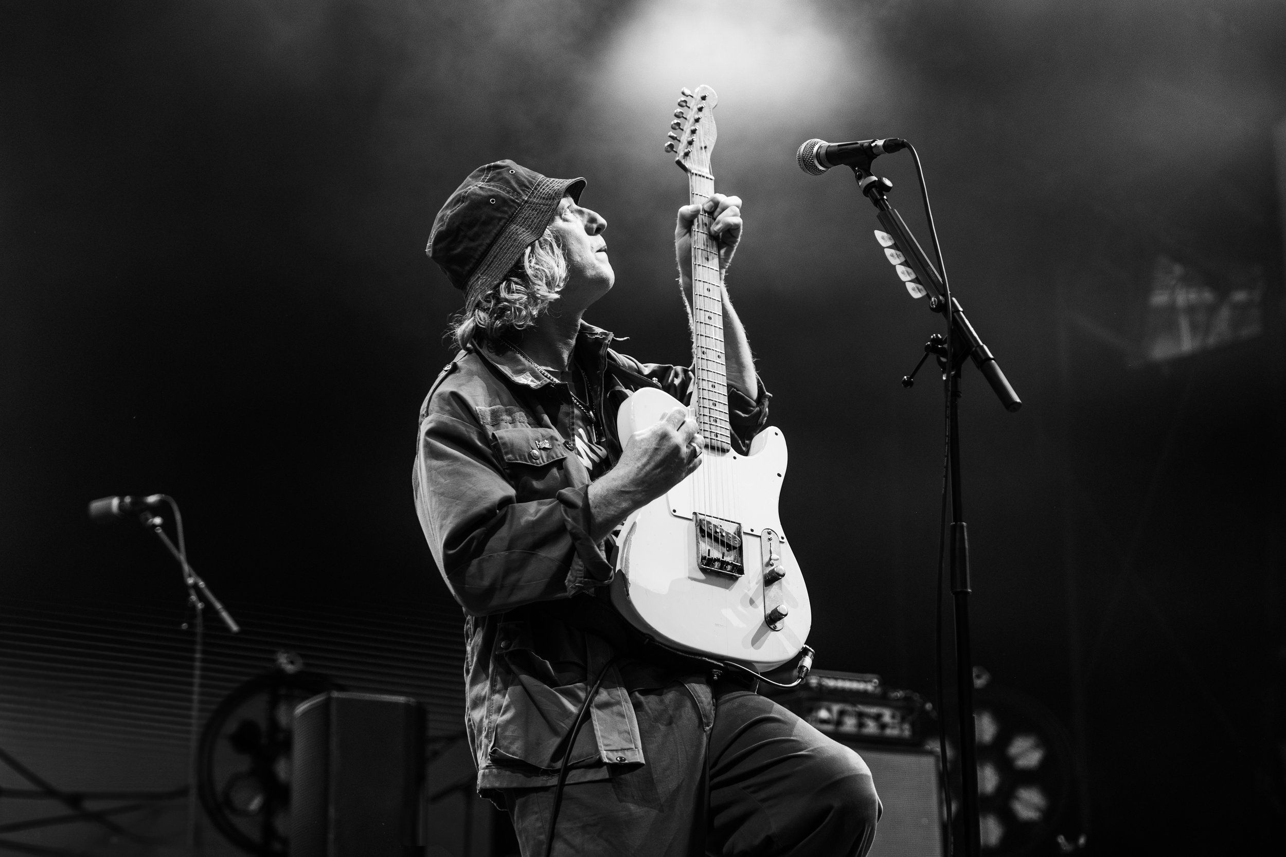 Black and white photo of a musician playing an electric guitar on stage with a microphone in front, wearing a hat and casual clothing.