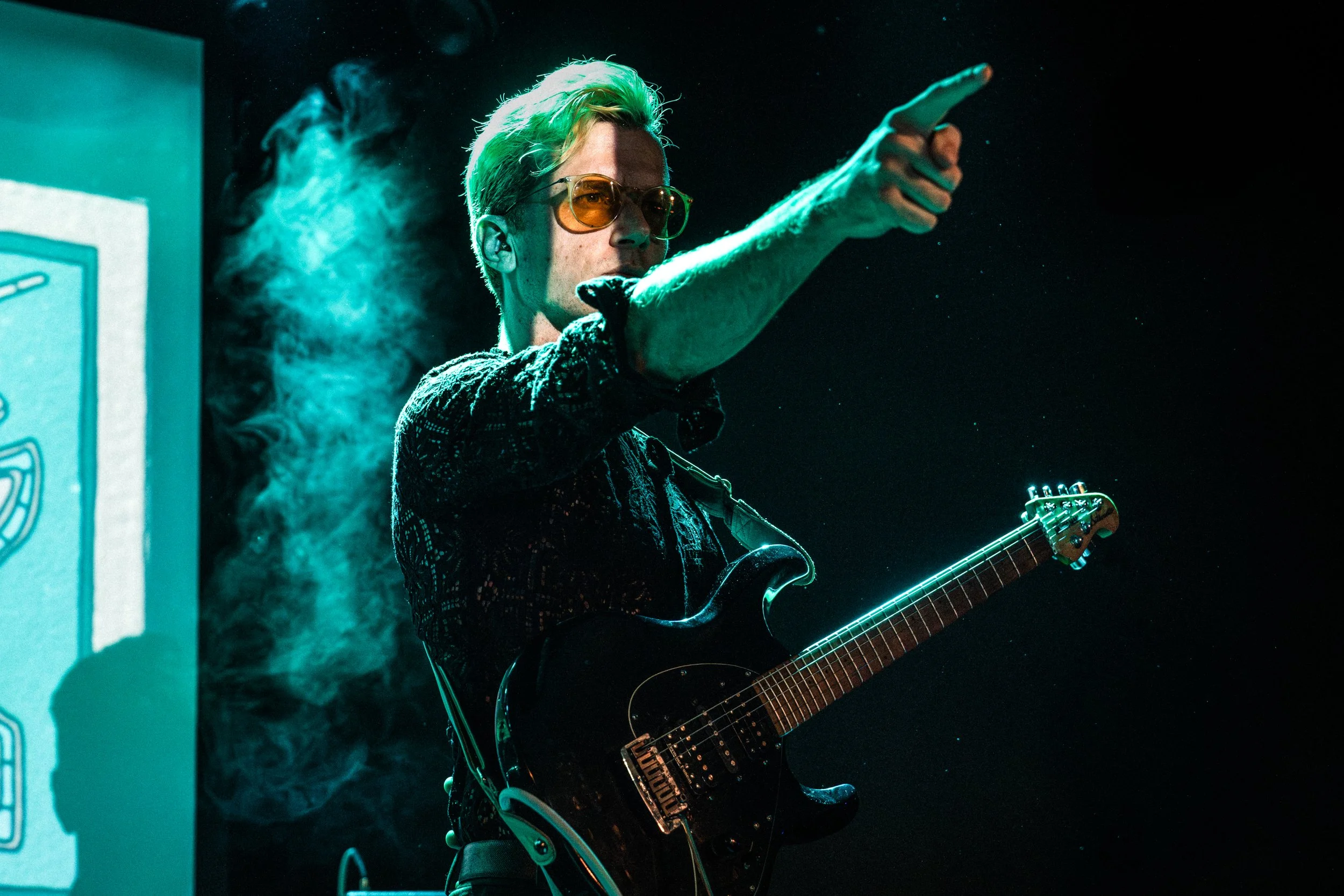 A man with blonde hair, sunglasses, and a black shirt is playing an electric guitar on stage. He is pointing with his right hand, and there is blue smoke and stage lighting around him.