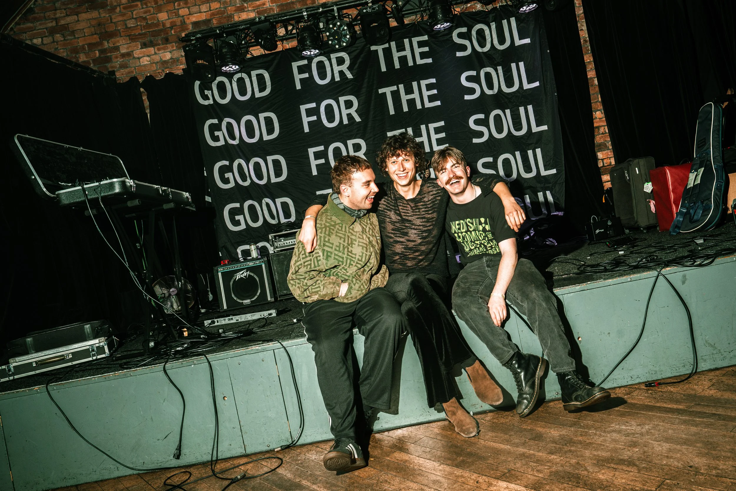 Three people sitting on a stage with musical equipment, smiling and hugging each other. Behind them is a black banner with the repeated phrase "Good for the Soul."
