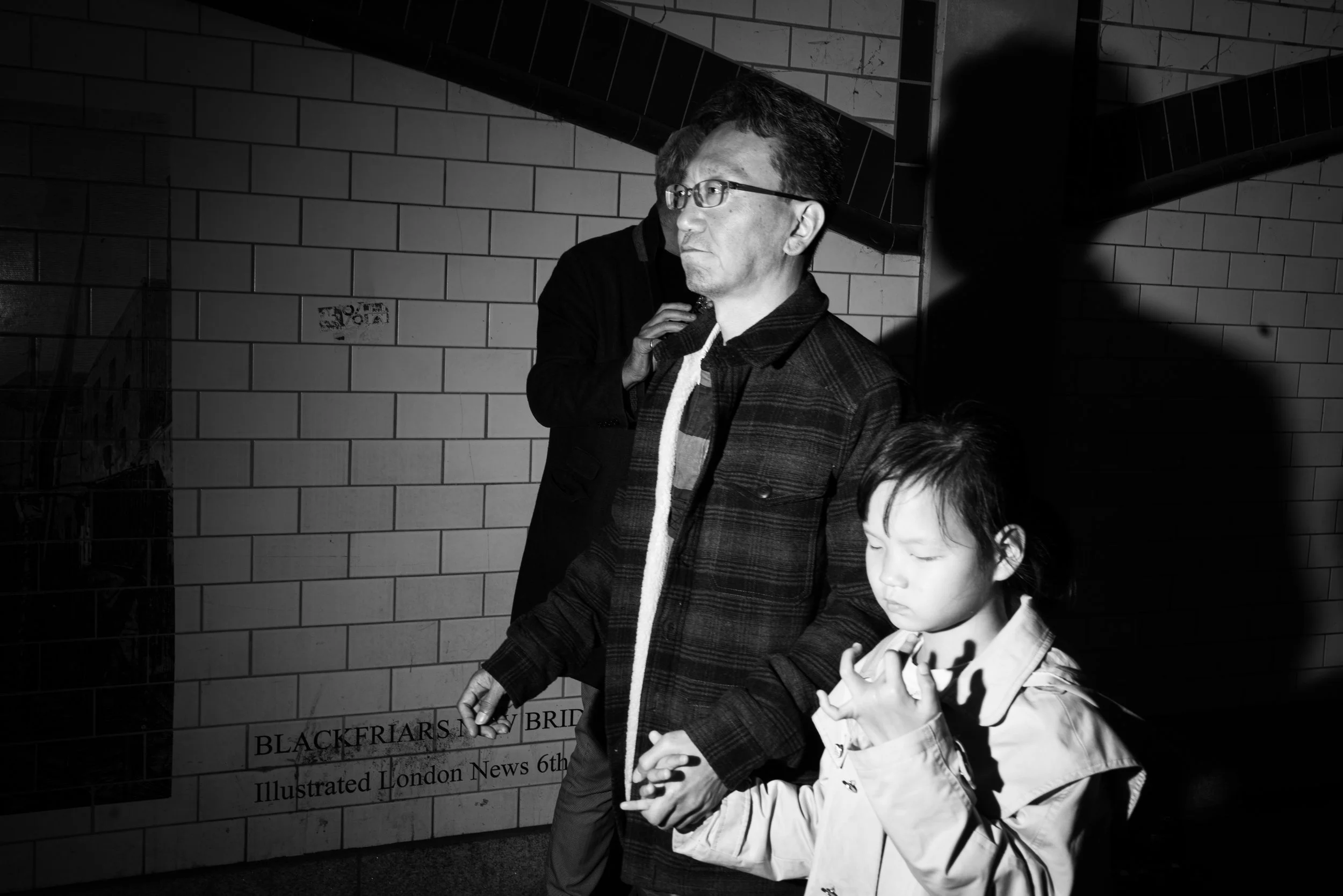 Black and white photo of an Asian girl with closed eyes and a contemplative expression standing in front of a brick wall, with a shadow cast by a person behind her, and a man wearing glasses and a dark jacket standing beside her.