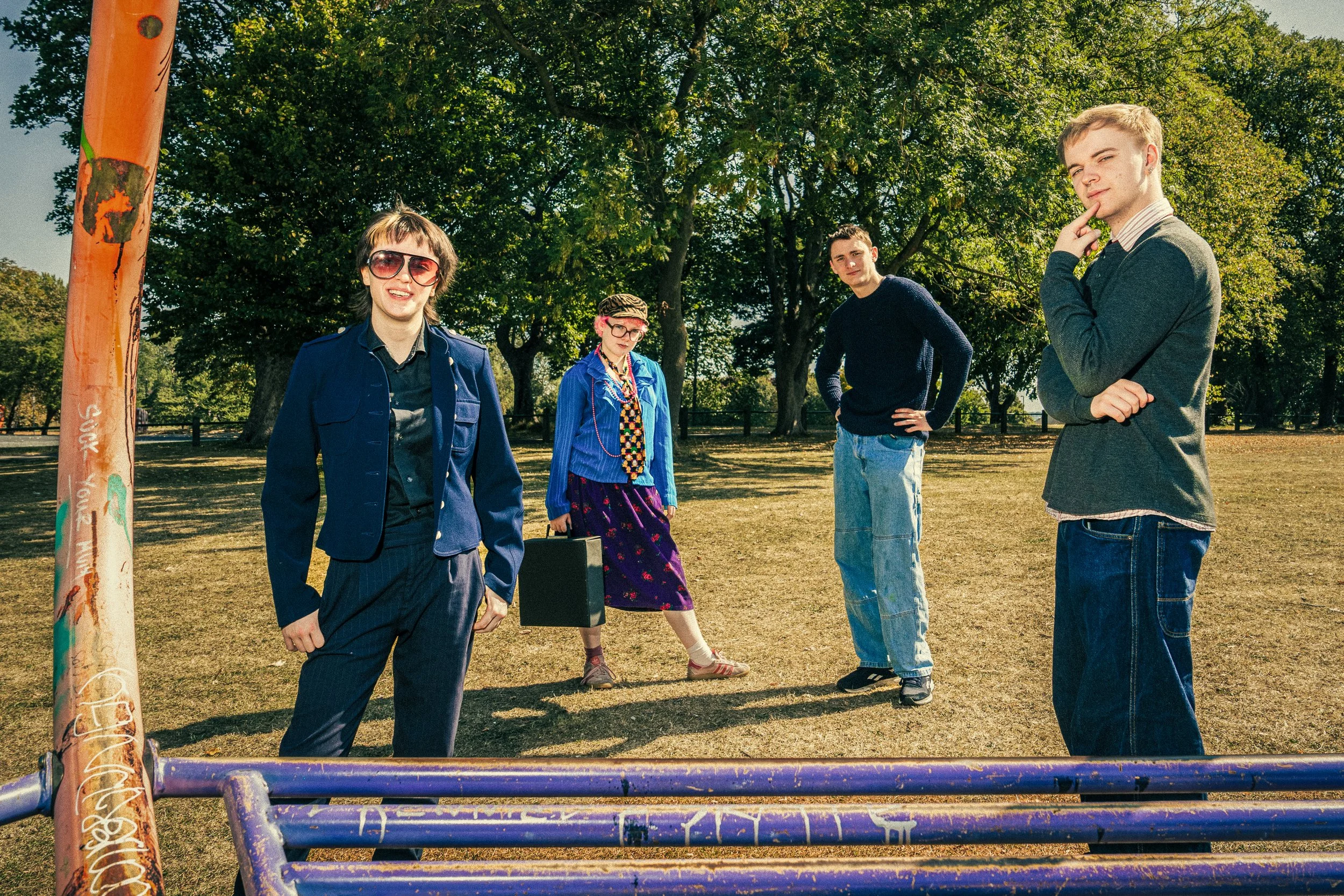Four young adults standing in an outdoor park on a sunny day, with two men and two women, dressed in casual and vintage-style clothing, some wearing glasses and one woman carrying a briefcase.