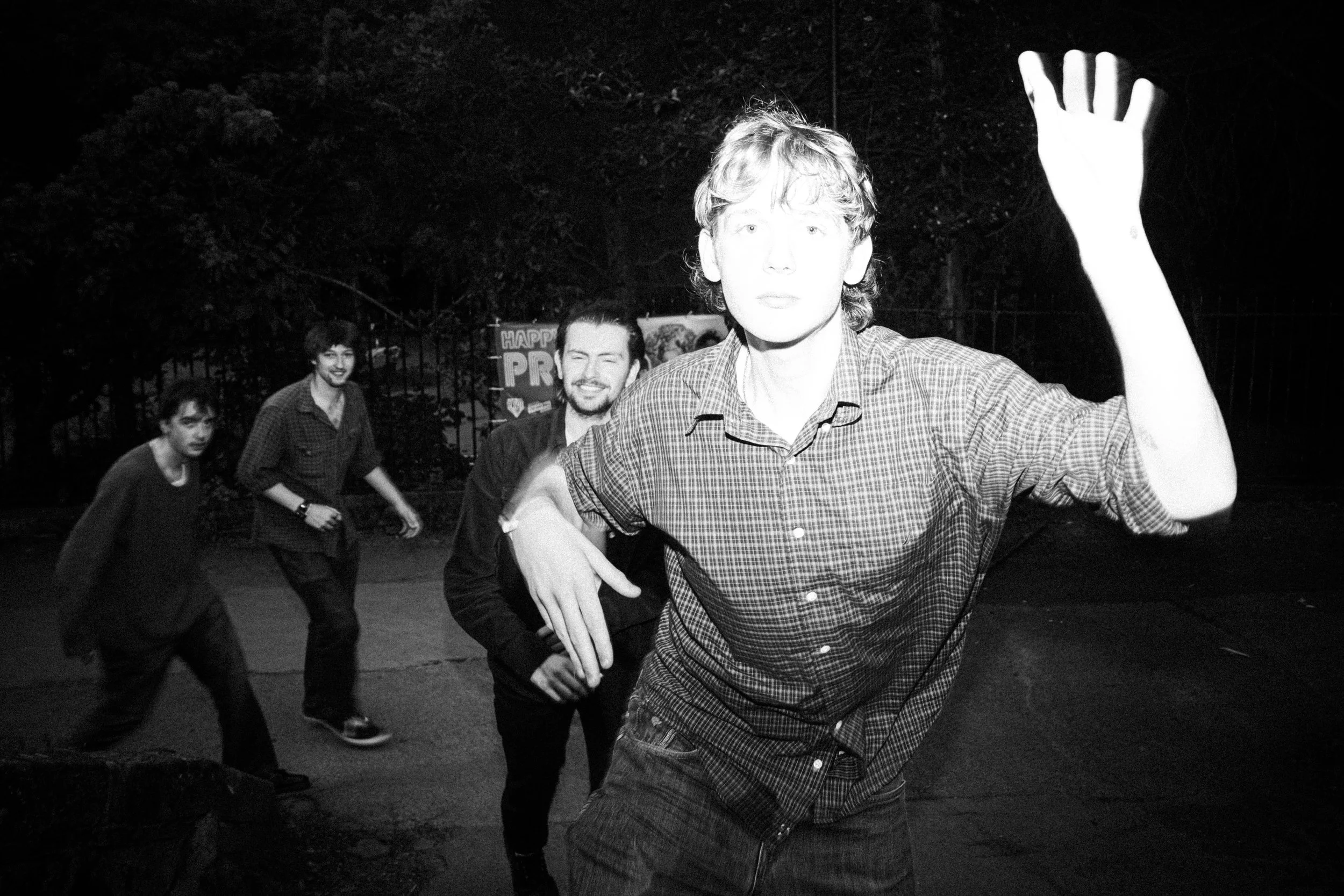 Black and white photo of four young men outdoors at night, with trees in the background. The man in the foreground is raising his hand and looking at the camera, while the other three are smiling and walking behind him.
