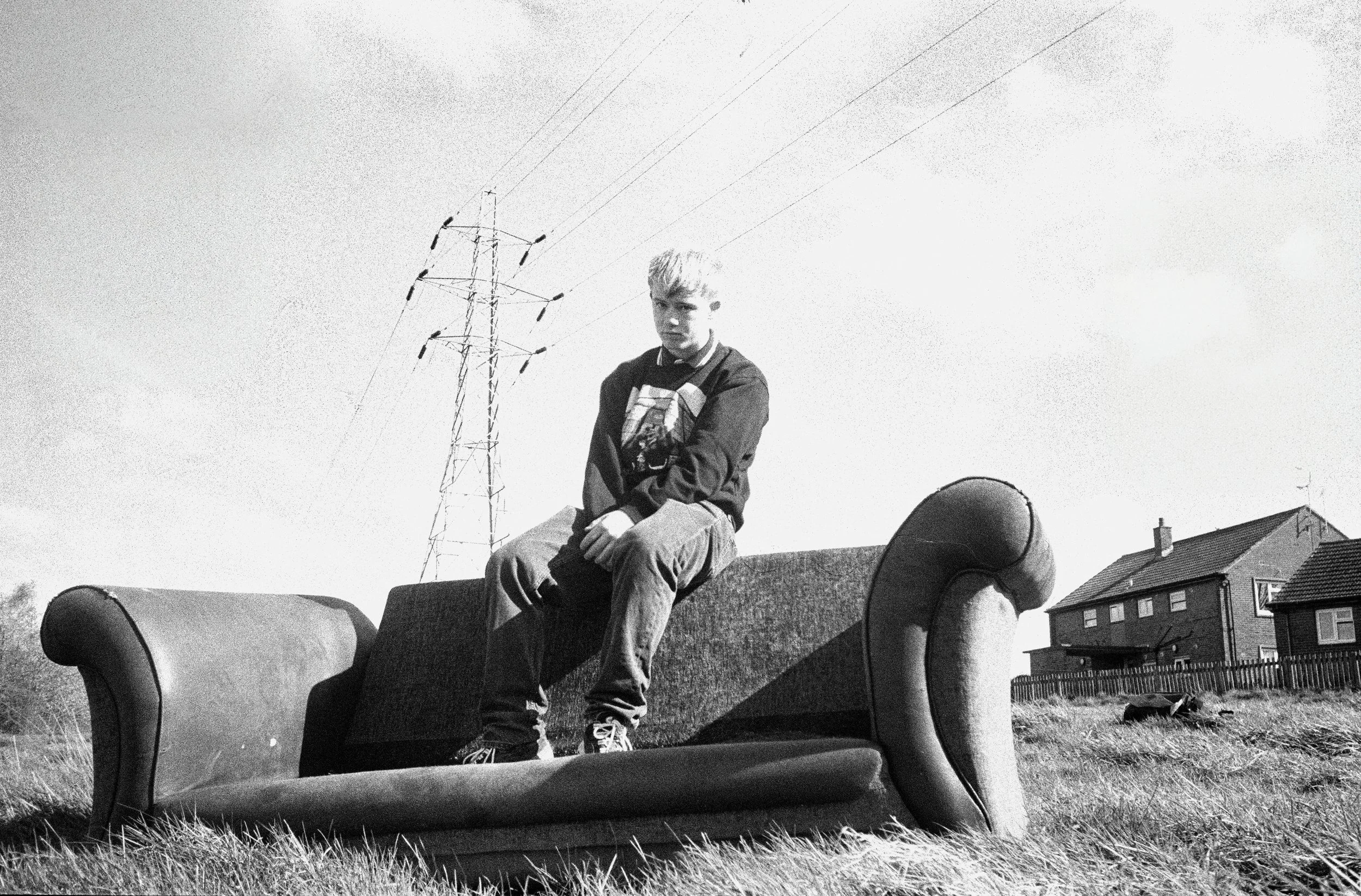 A young boy sitting on a vintage sofa outdoors on grass, with a residential neighborhood and power lines in the background, black and white photo.