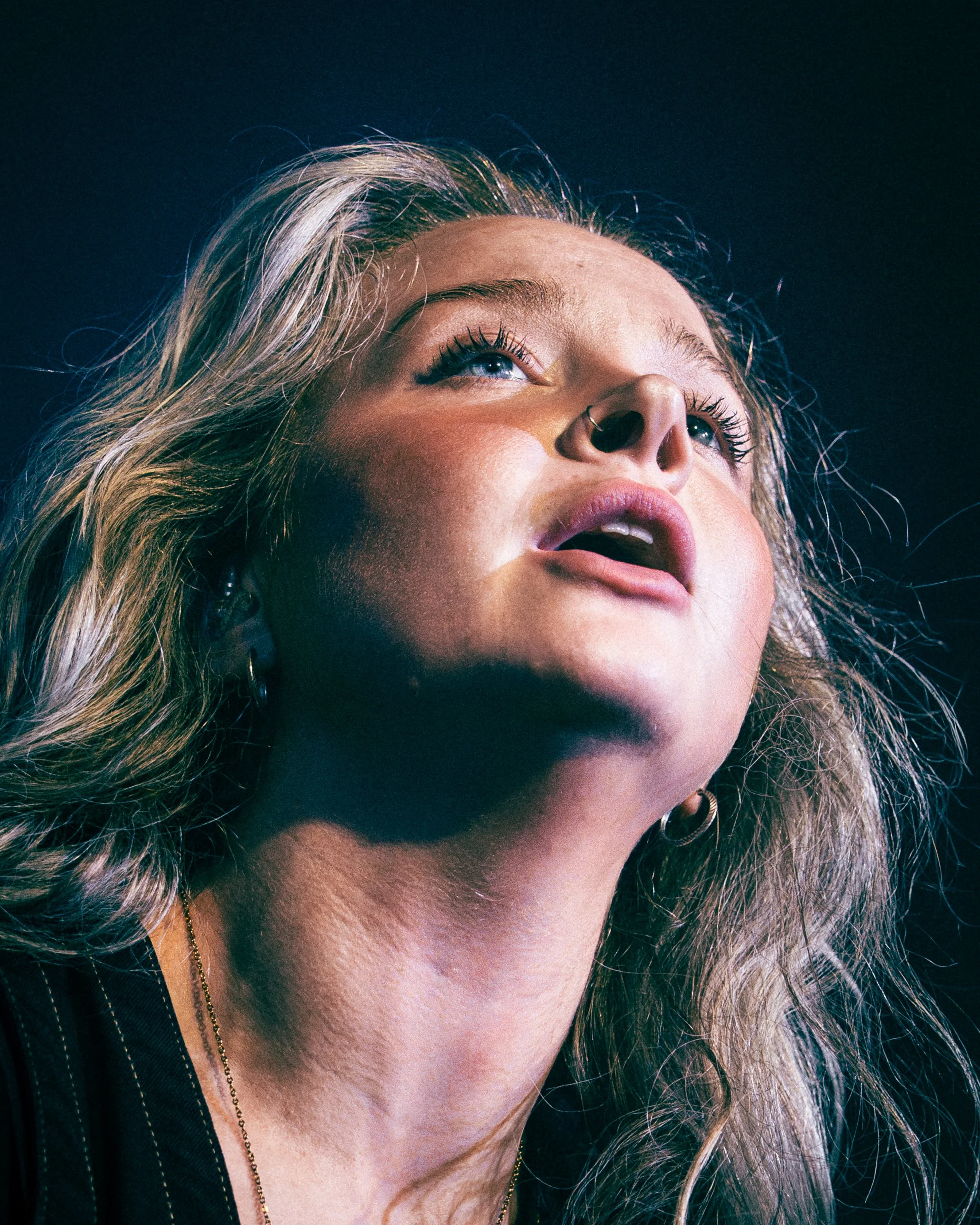 Close-up of a woman with blonde curly hair and a septum piercing looking upward.
