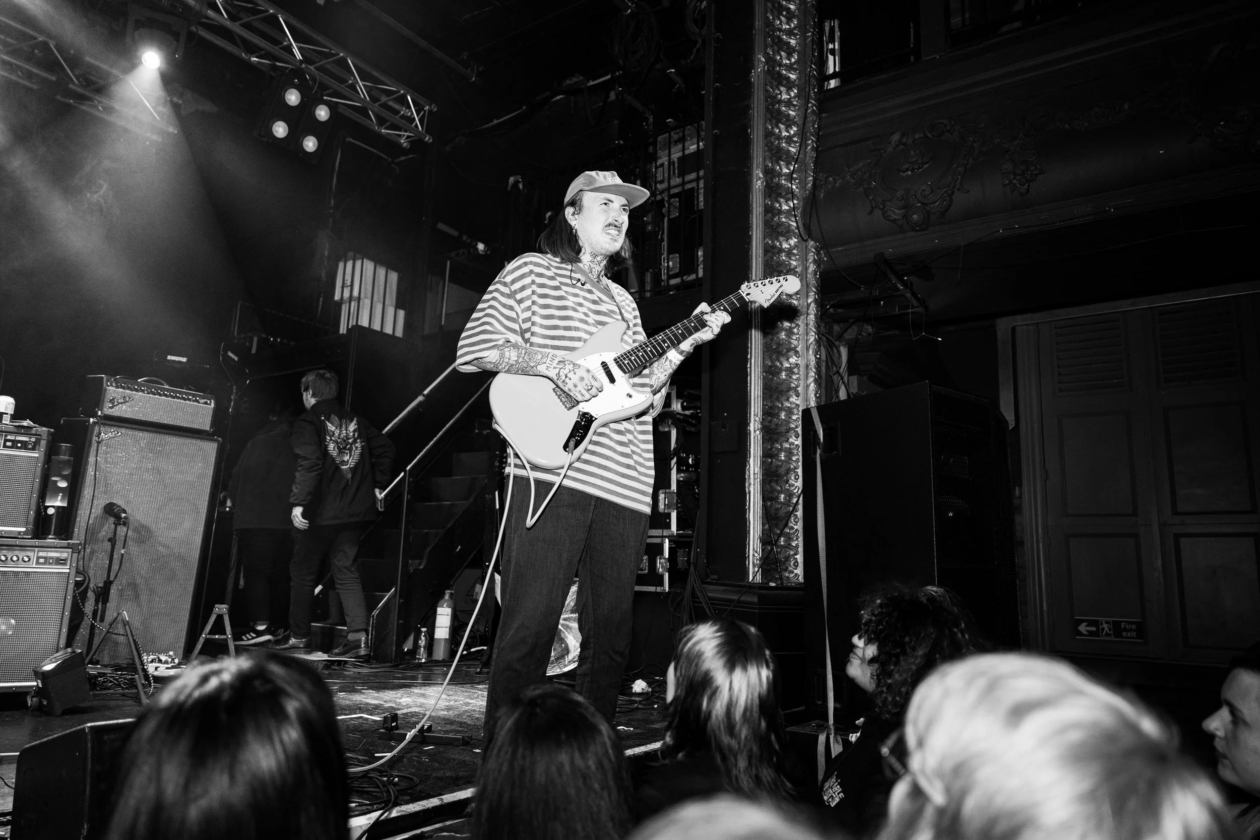 A musician on stage playing an electric guitar, wearing a striped shirt and baseball cap, with audience members watching nearby.
