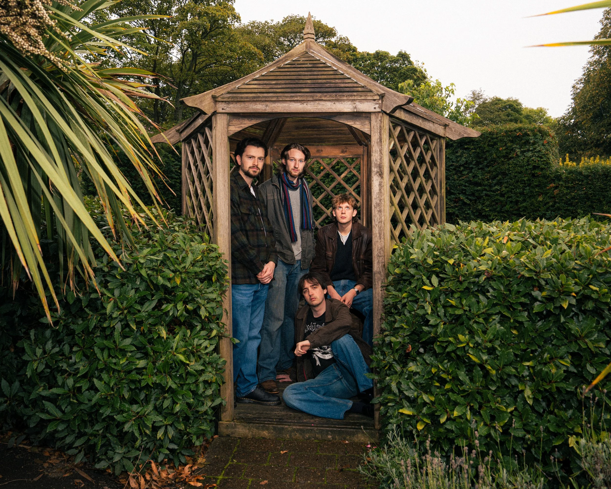 Five young men with dark hair and casual clothing, some with jackets, pose in and around a small wooden garden shed surrounded by lush green bushes and trees during daytime.