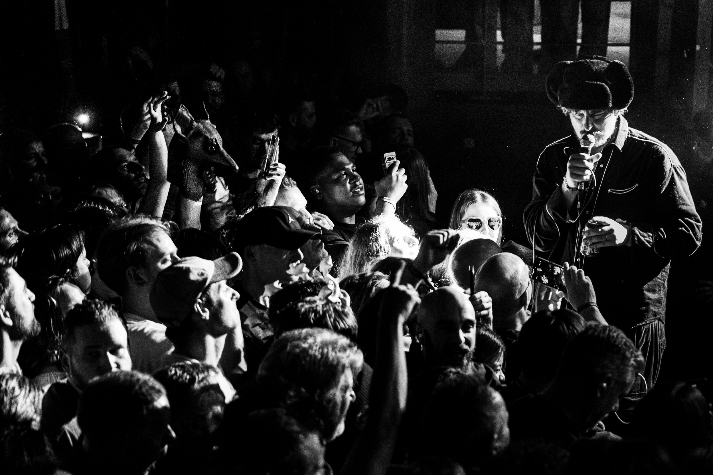 A black and white photo of a male performer on stage holding a microphone, surrounded by a crowd of people taking photos and videos with their phones, some with raised fists and sunglasses, in a lively concert environment.