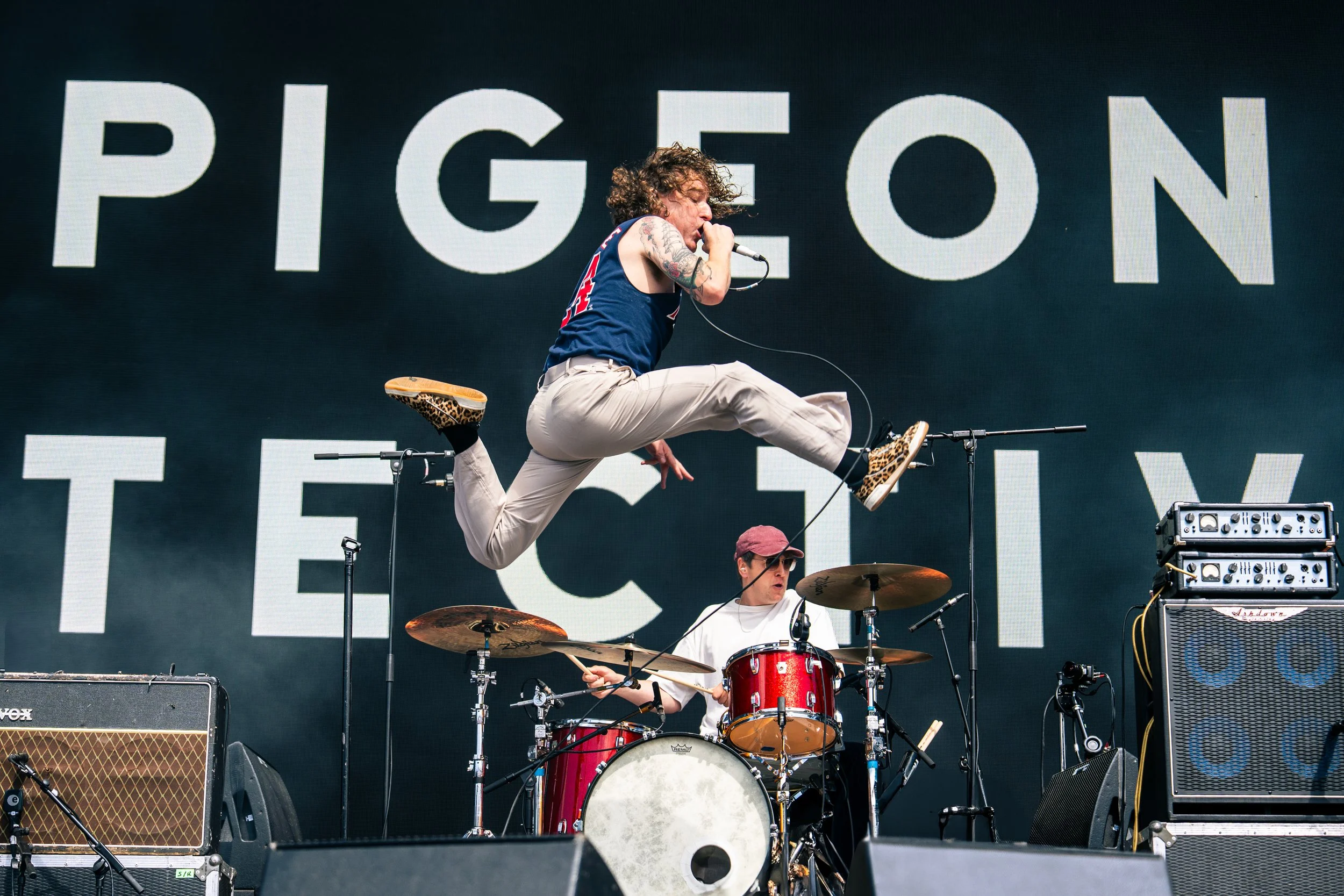 Band performing live at a concert with a singer jumping in the air and a drummer playing behind on stage.