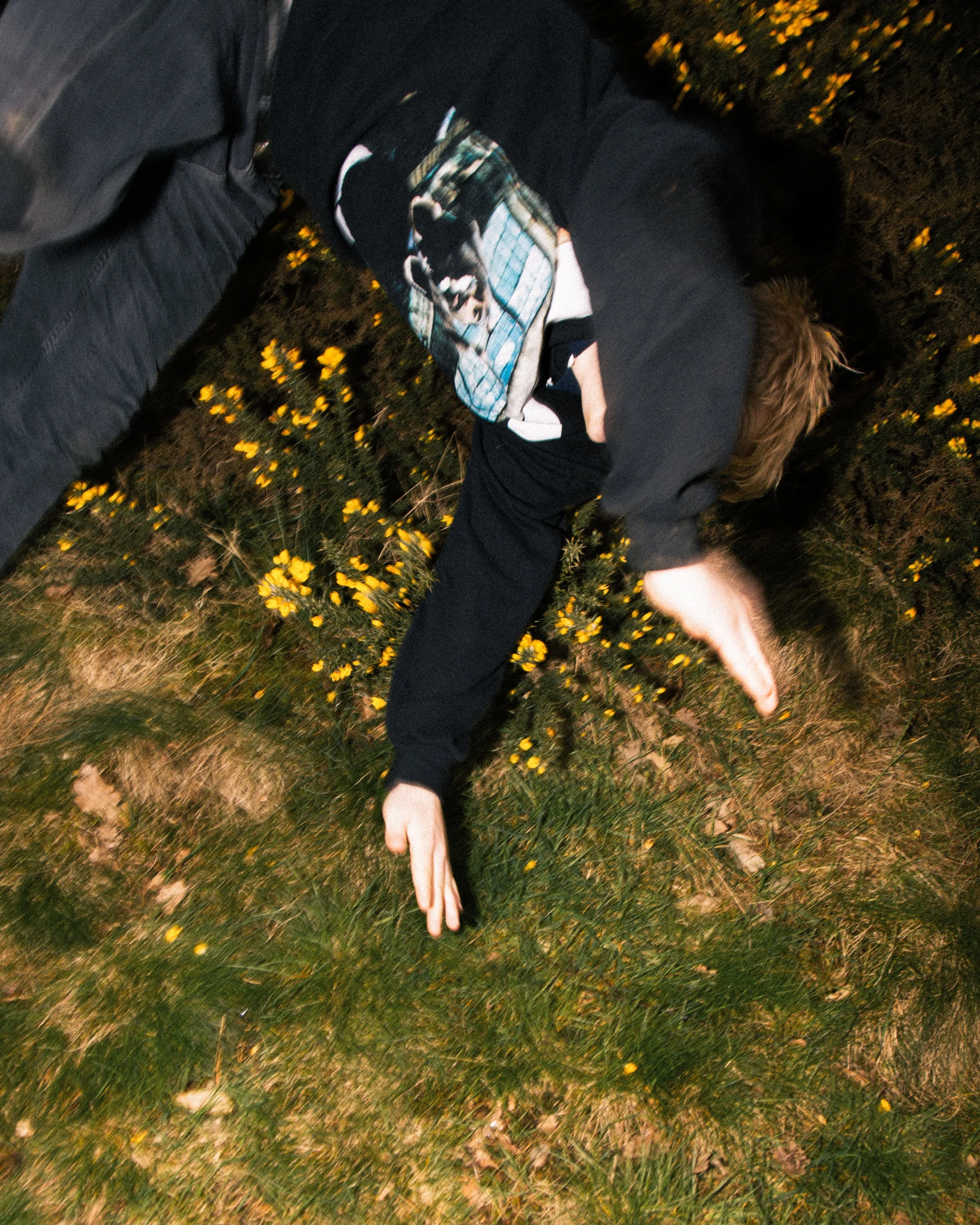 Person laying on the ground in a grassy area with yellow flowers, wearing a black hoodie and dark pants, at night.