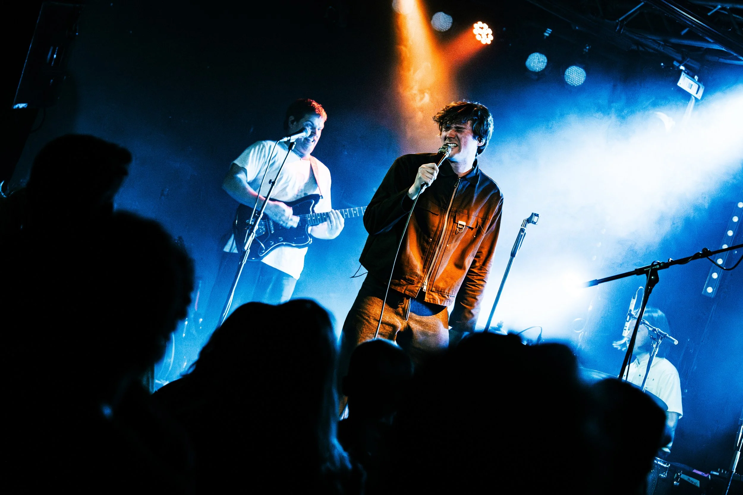 Two musicians perform on stage with blue and orange lighting, one singing into a microphone and the other playing an electric guitar, with a crowd in the foreground.