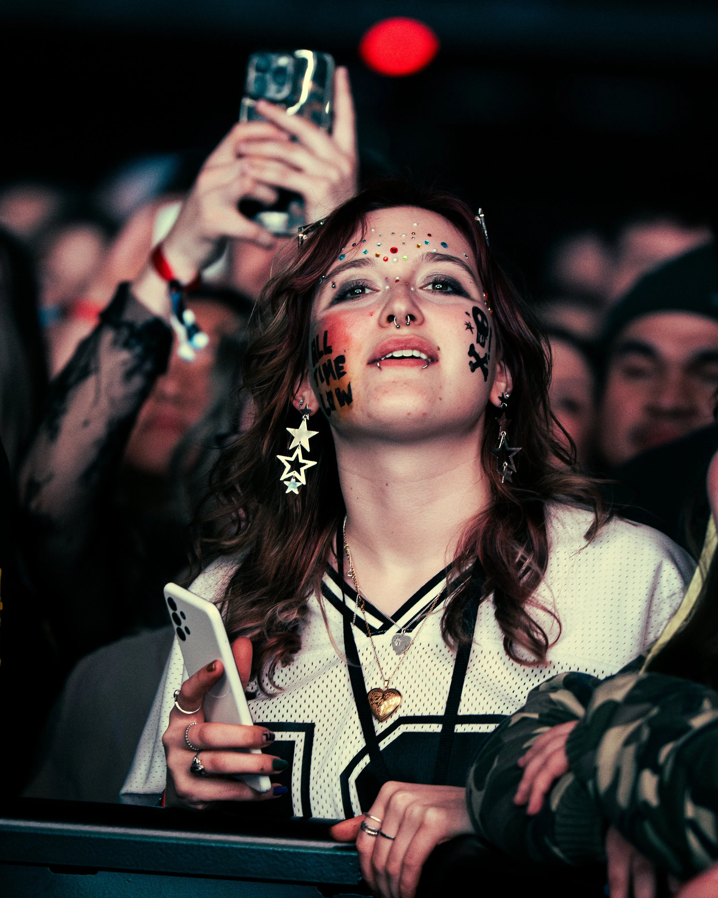 A young woman with face decorations and earrings looking up at a concert, holding a smartphone, with a crowd in the background.