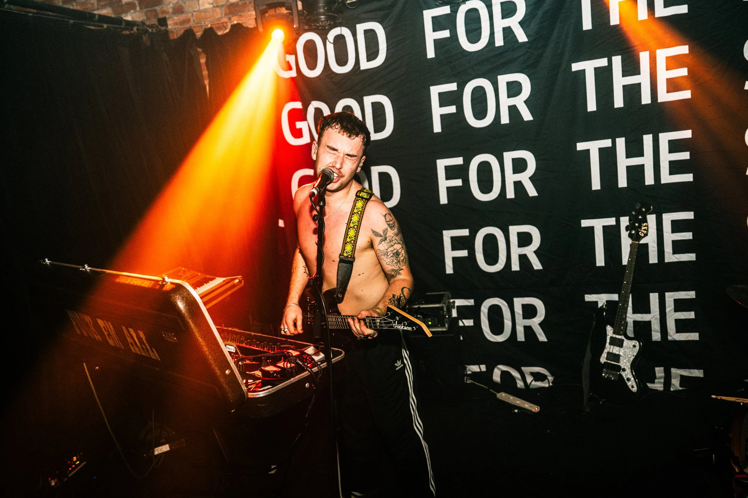 A shirtless male musician with tattoos, singing into a microphone, playing an electric guitar, in a dark performance space with orange stage lights and a black backdrop with white text that reads 'GOOD FOR THE' repeatedly.