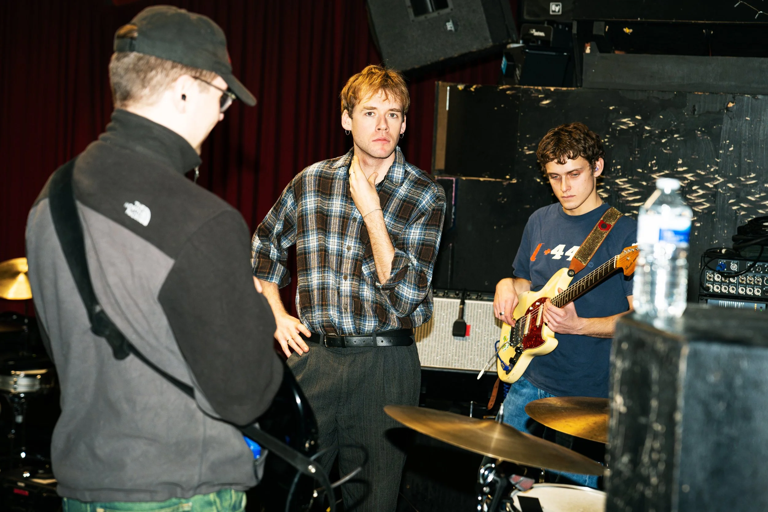Three young men in a music rehearsal space, one with a guitar, one with a bass, and one in a plaid shirt, engaged in conversation.
