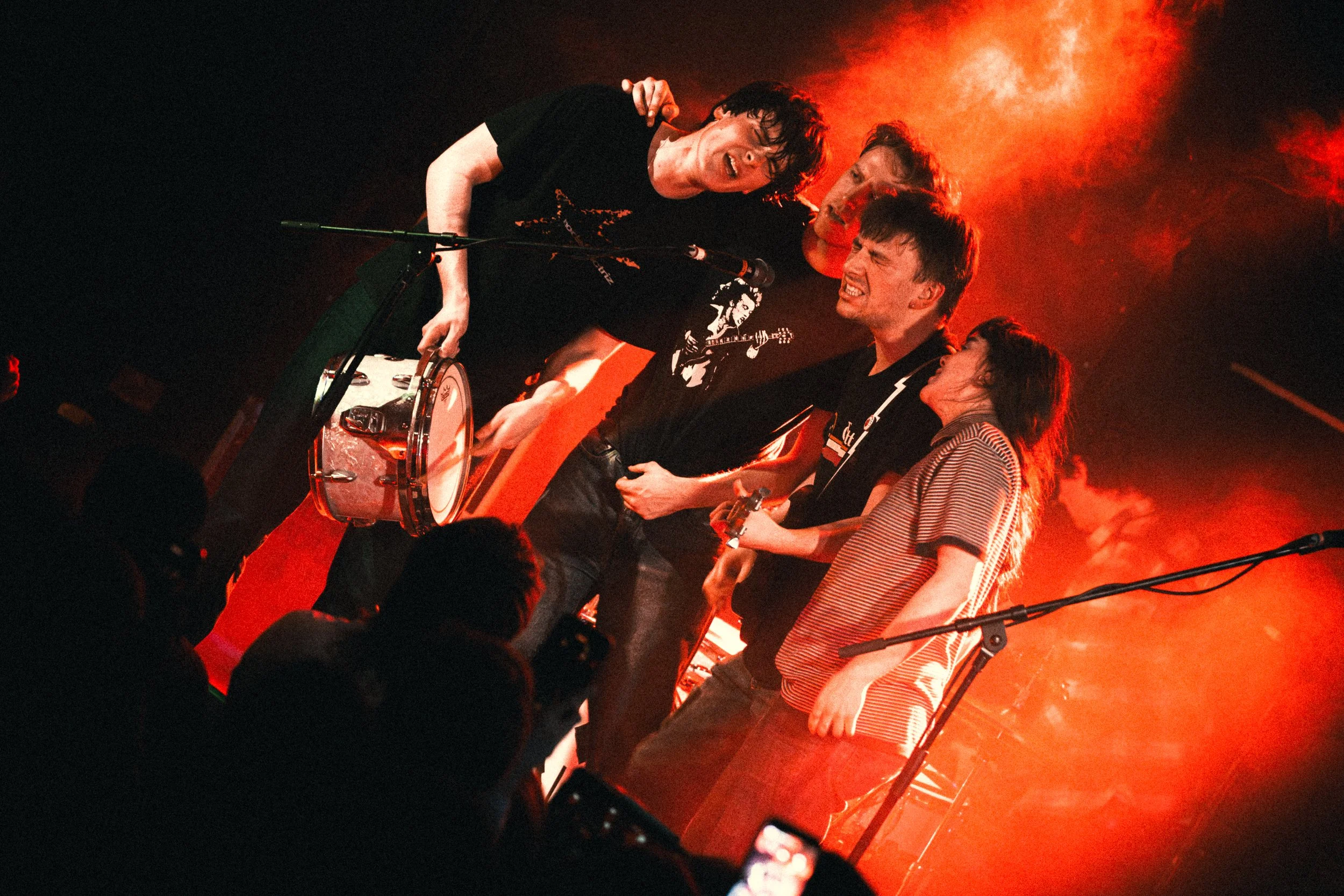 Four band members performing on stage with red lighting and smoke, holding instruments, with an audience in front.