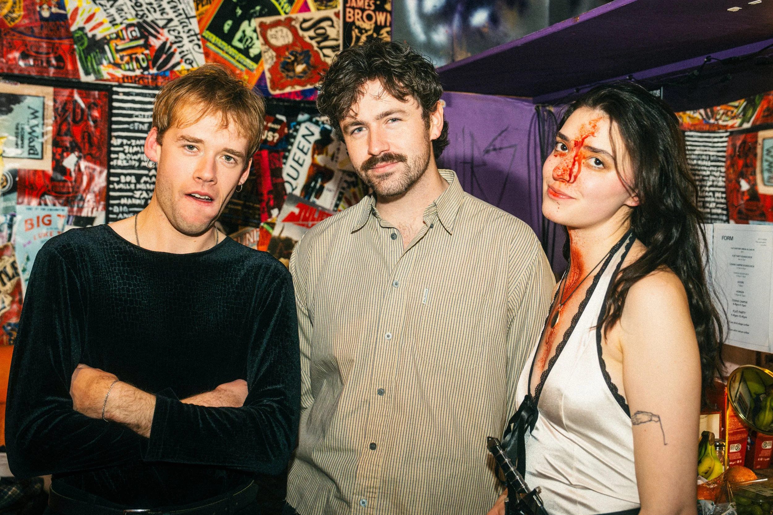 Three people standing in front of a colorful, collage-style wall with various posters and paper items, at an indoor event.