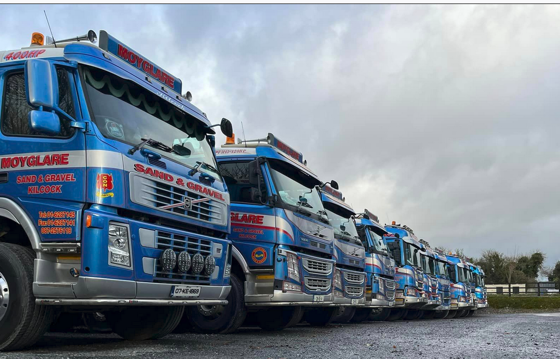 A line of blue trucks with red and white text reading 'Moyglare' and 'Sand & Gravel Kilcock' parked outdoors on a cloudy day.