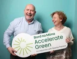 A smiling man and woman holding a sign promoting Accelrate Green, a partnership with Bord na Móna, against a teal background.