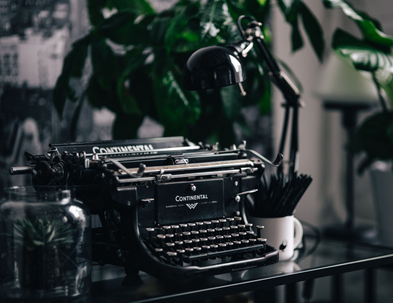 A vintage black typewriter with the brand name 'Continental' on it, placed on a black surface in front of leafy green houseplants, with a small jar and a white cup holding black straws nearby.