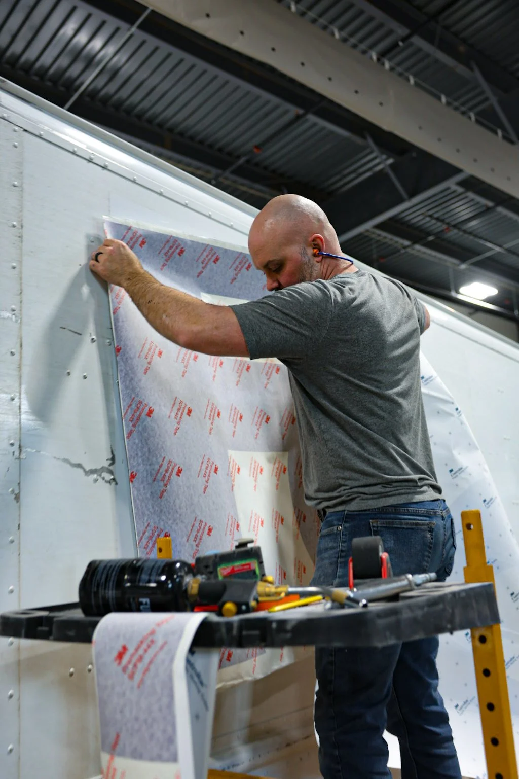 Brian Barenbrock, Collision Repair Foreman at Optimus Group (Optimus Body Shop), installing stickers on a vehicle in a body shop setting, with tools on a work table nearby.