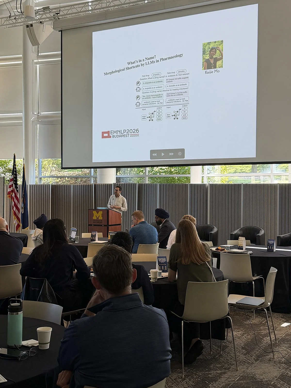  Dr. Byron Wallace, one of the day’s two keynote speakers, presents at the podium during the AI &amp; Health Symposium. Behind Dr. Wallace, the projector screen displays a slide titled, “What’s in a Name? Morphological Shortcuts by Large Language Mod