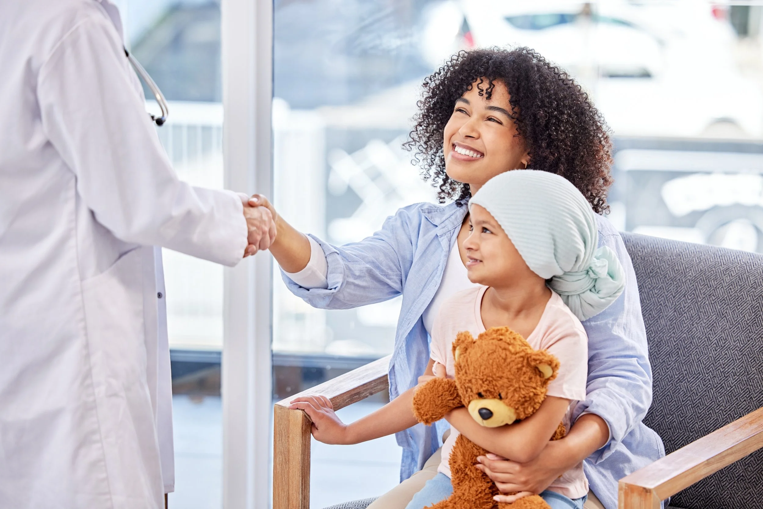 A mother sits with her child in her lap in the waiting room of a healthcare facility. The top of the child's head is covered in a bandana, suggesting hair loss due to illness. The mother is smiling and shaking the hand of a doctor.