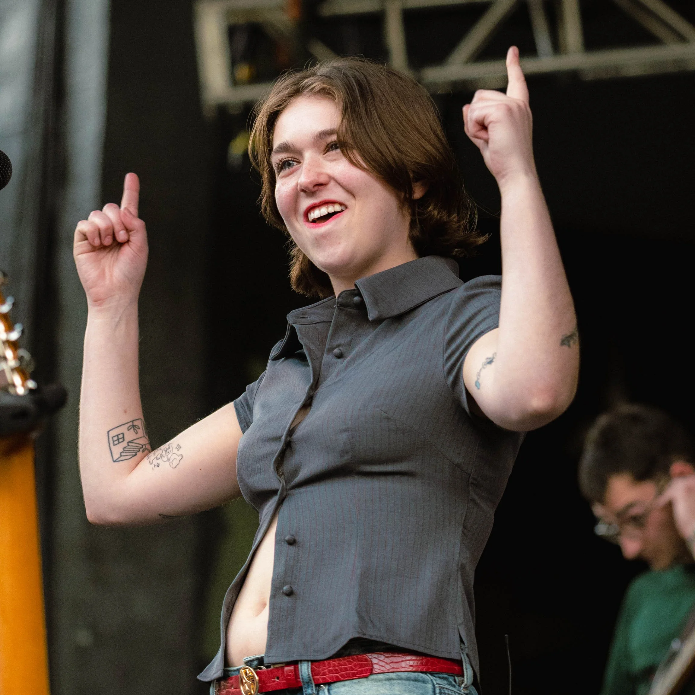 A young woman with short brown hair smiling and raising her arms, wearing a dark gray shirt with rolled sleeves, jeans, and a red belt.
