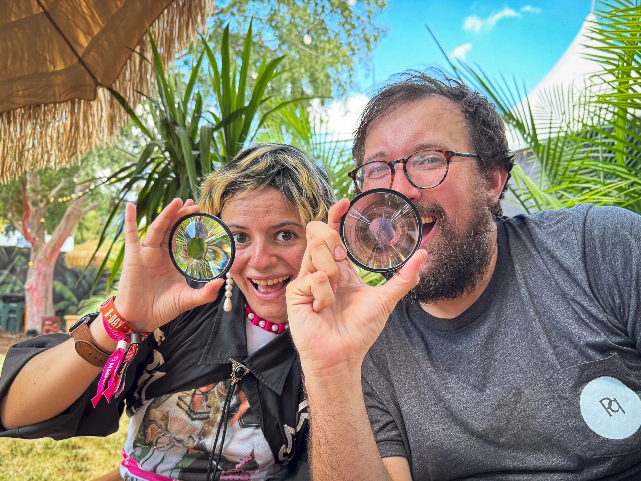 A man and a woman smiling and holding up magnifying glasses with colorful reflections, outdoors with trees, plants, and a bright blue sky in the background.