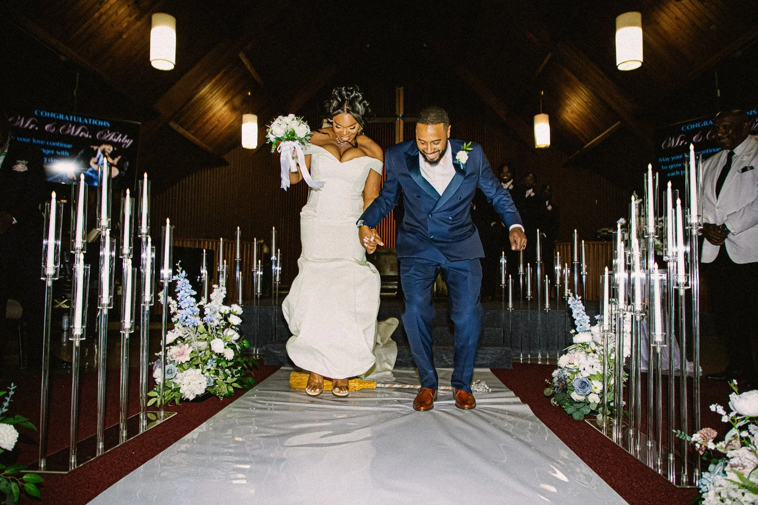 A newlywed couple happily walks down the aisle holding hands after their wedding ceremony, surrounded by floral decorations and candle holders, with wedding guests in the background.