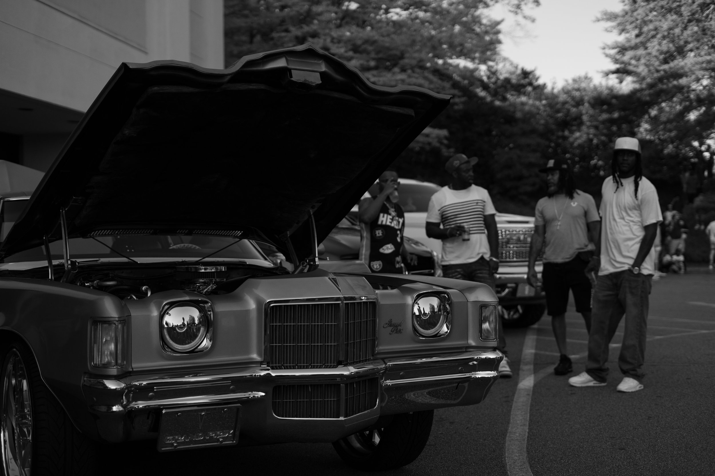 A vintage car with its hood open at a car show, with four men standing nearby, some holding drinks, and trees in the background.