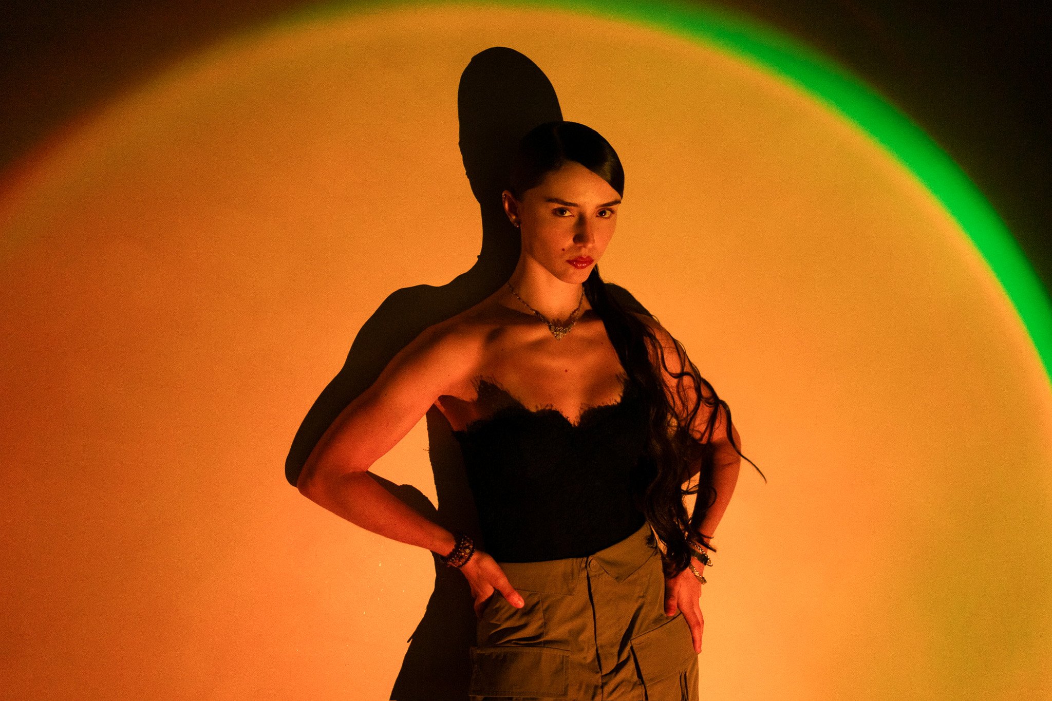 A young woman with long dark hair in a braid, wearing a black strapless top and khaki cargo pants, stands against a yellow-orange background with a green arc on the upper right. She is posing with hands on her hips, looking at the camera with a serious expression, and casting a distinct shadow.