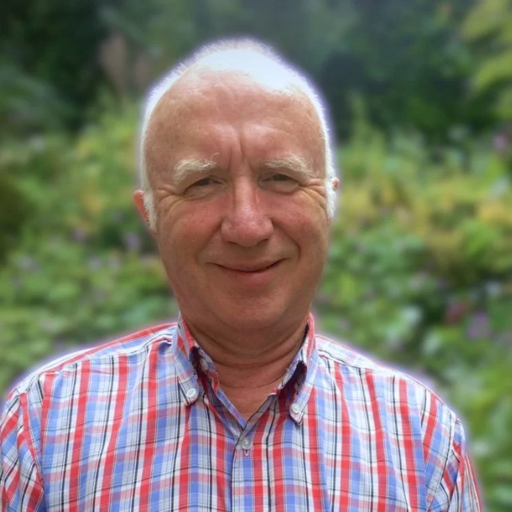 Smiling older man with short white hair wearing a red and blue plaid shirt, standing outside with greenery in the background.