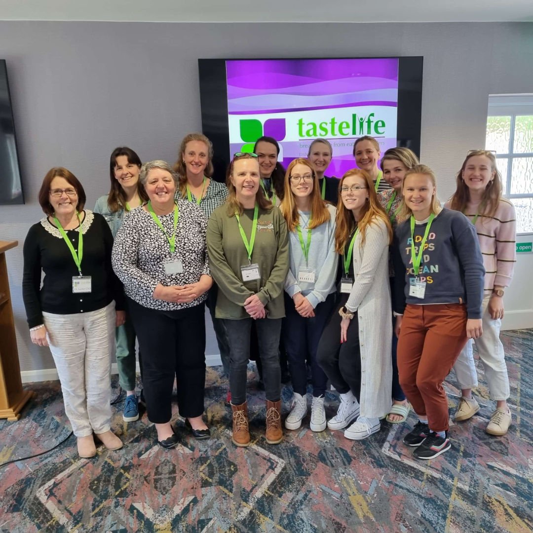 Thirteen smiling people stand indoors in front of a tastelife presentation screen, all wearing name badges and green lanyards.