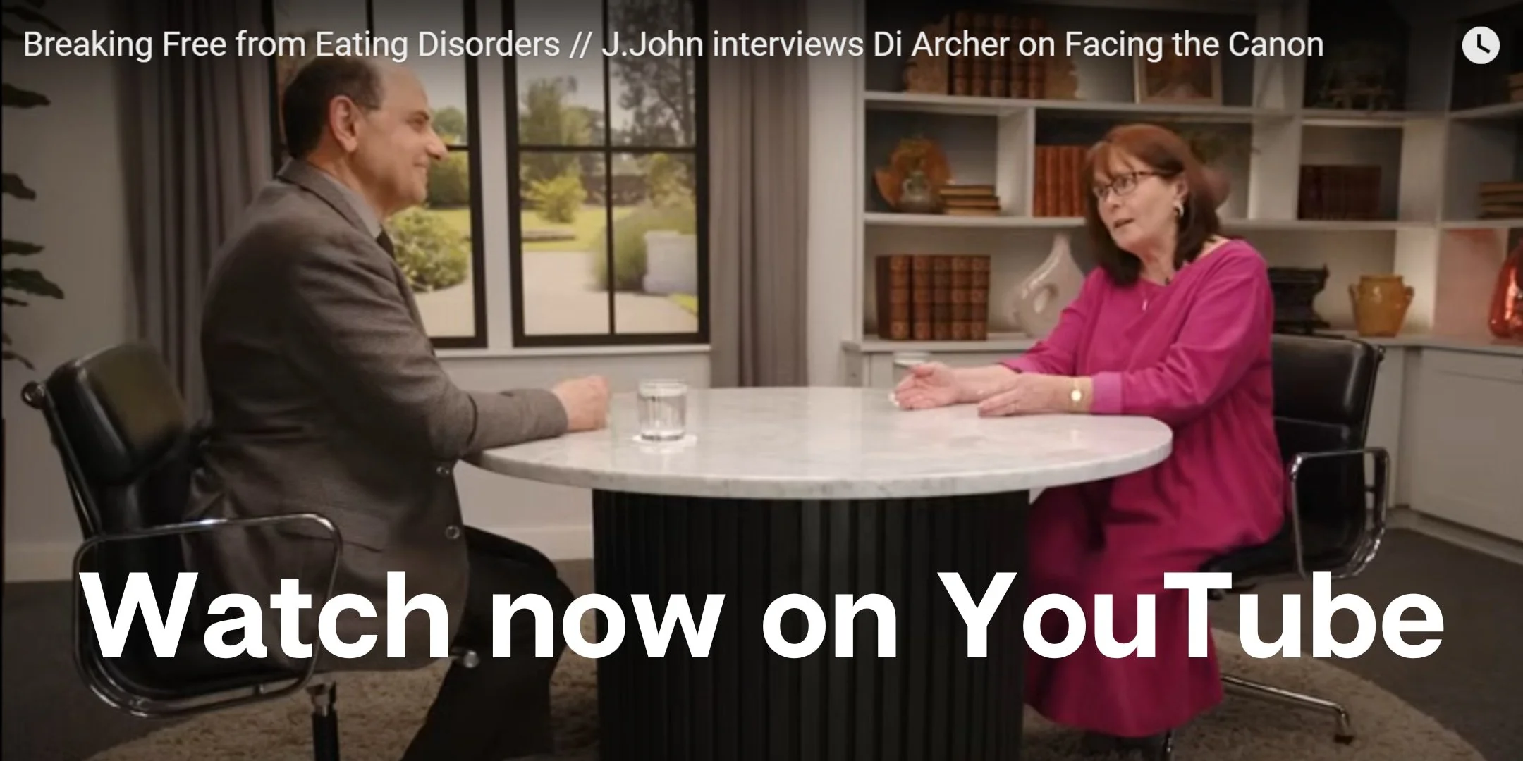 An interview scene with a man in a gray suit and a woman in a pink blouse sitting across from each other at a white round table in a room with large windows and a bookshelf.