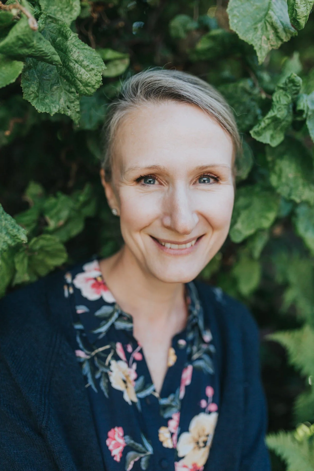A woman with short blonde hair and blue eyes smiling, surrounded by green leaves.