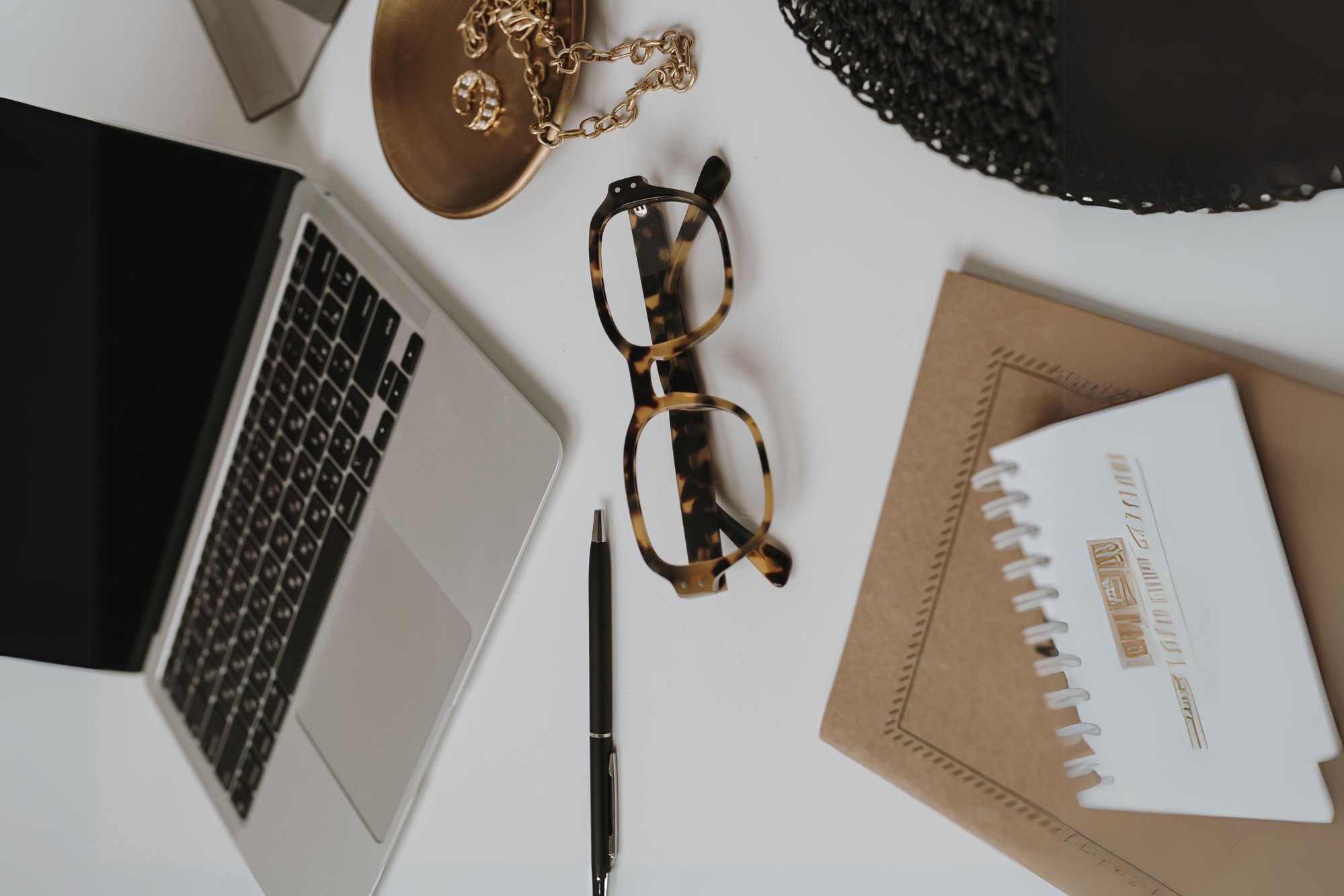 A laptop, a pair of tortoise shell glasses, a black pen, a spiral notebook, a brown notebook, gold jewelry including a chain and earrings, and a black woven hat or basket on a white desk.