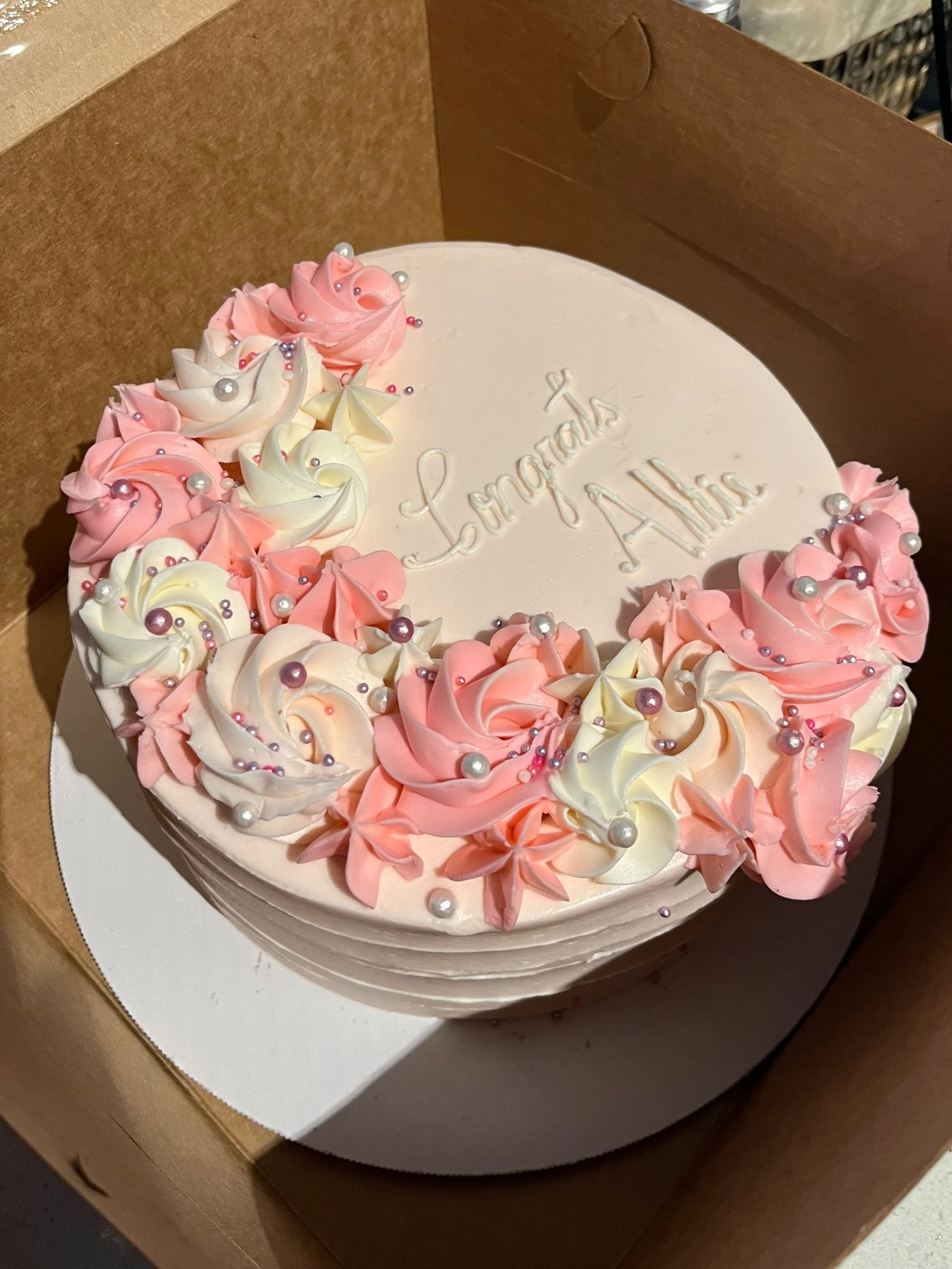 A birthday cake with white icing, decorated with pink and white buttercream flowers and small pearl-like edible beads, inside a cardboard box. The cake has the inscription 'Laraspiki's ALESS' on top.