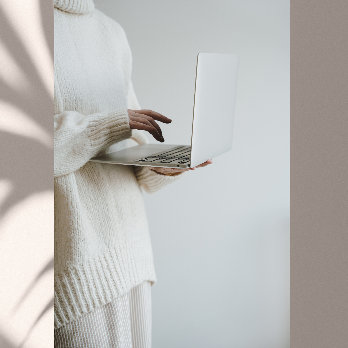 Person wearing cream-colored sweater holding a silver laptop in front of a plain, light gray background.