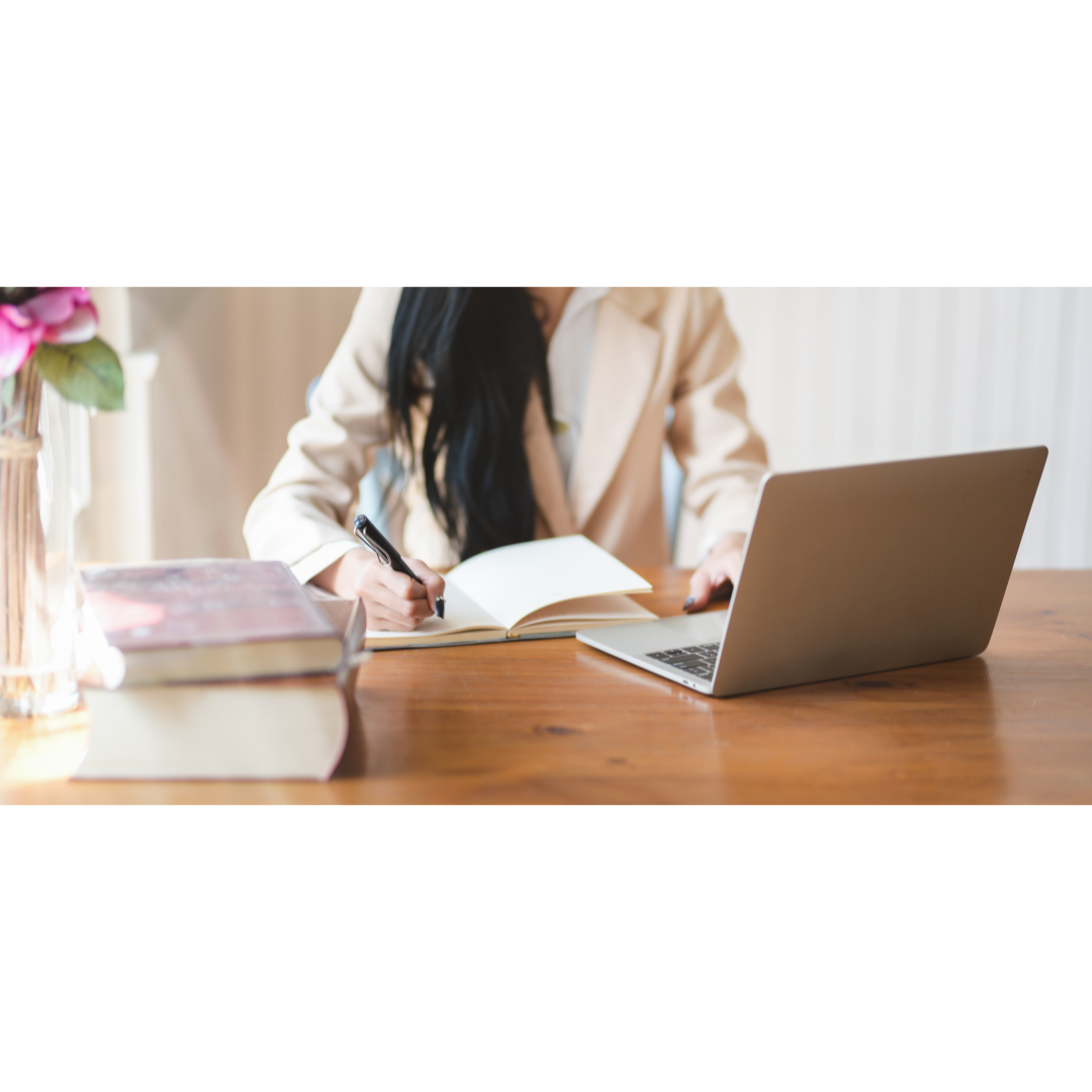 Person sitting at a wooden table, writing in a notebook with a pen, with a laptop and a stack of books nearby.
