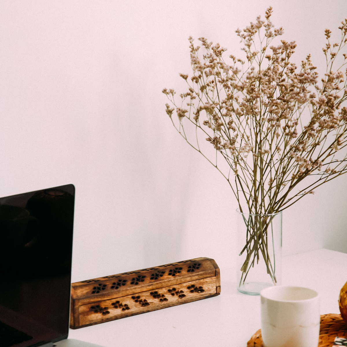 A white desk with a bouquet of pink flowers in a clear glass vase, a white cup, a wooden rectangular box with black carved patterns, part of a laptop on the left, and a woven placemat with a croissant on it.