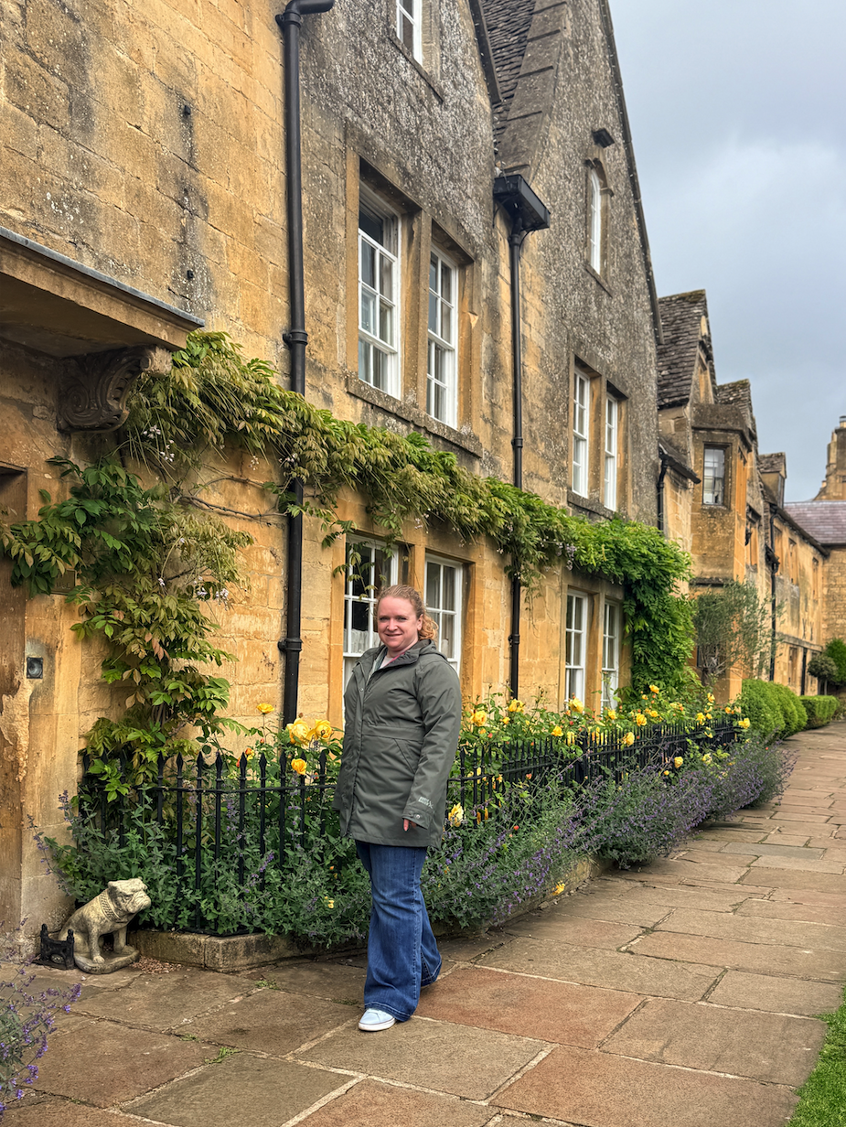 Anna Fishman of Olegana Travel Boutique standing on stone sidewalk outside honey-colored Cotswolds stone cottages in England with garden flowers.