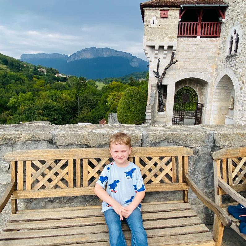 Anna and Oleg, young child sitting on a wooden bench at a stone castle courtyard with the Dolomite mountain range in the background during spring.