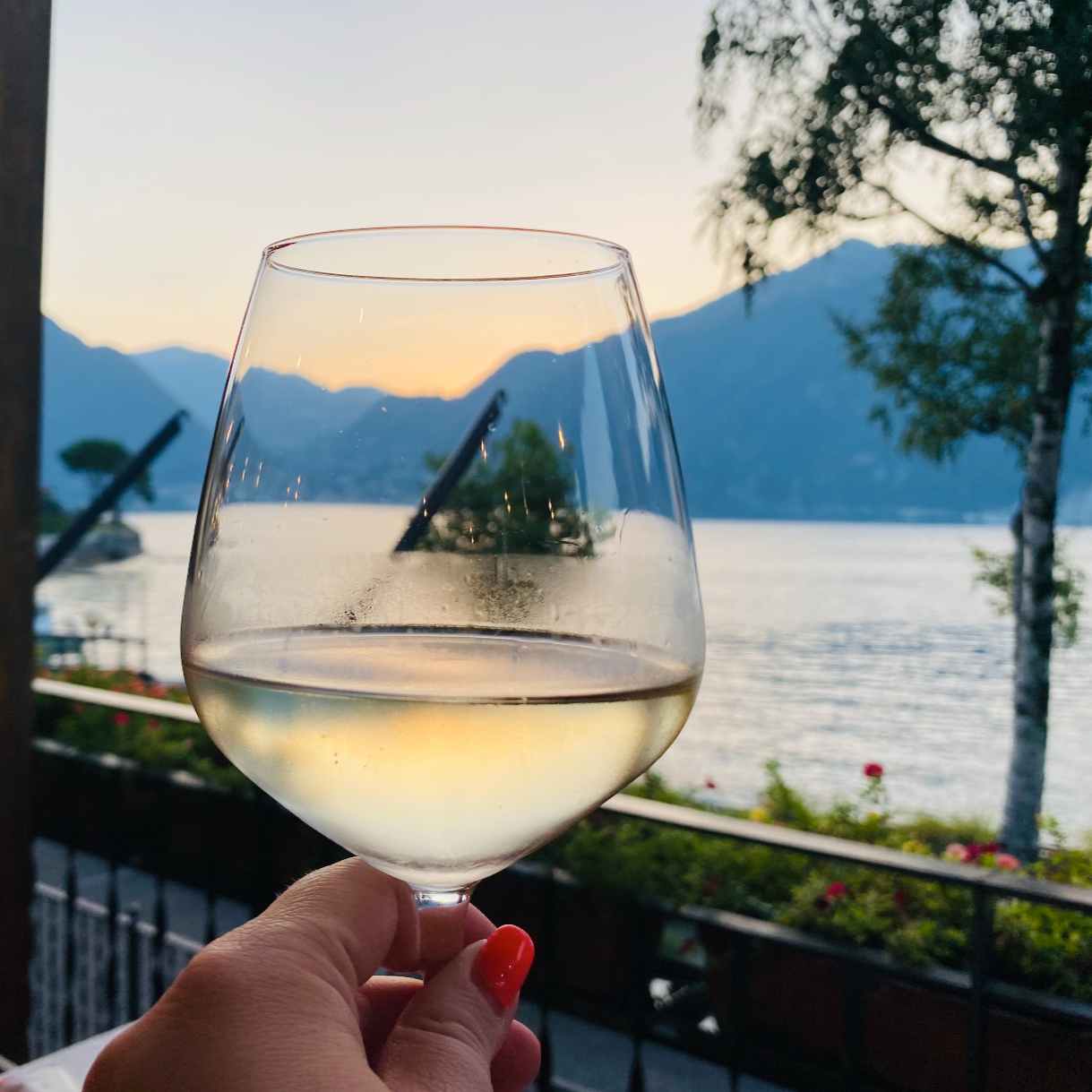 Anna Fishman holding a glass of chilled white wine at an outdoor patio during sunset with scenic lake and mountain views in the background