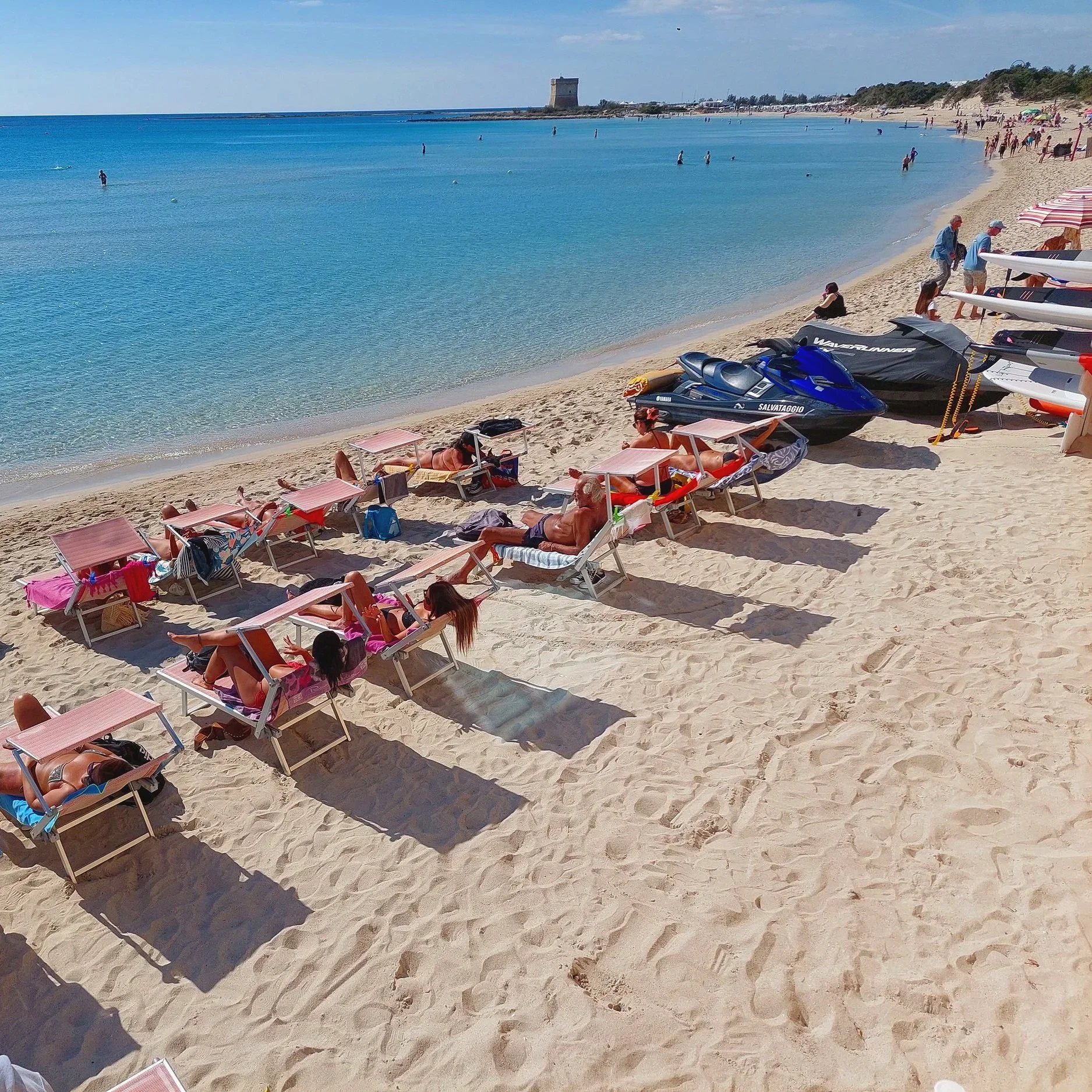 Porto Cesareo, Puglia, Italy, featuring a lively sandy beach with sunbathers relaxing along the shore and jet skis cruising through the turquoise waters.