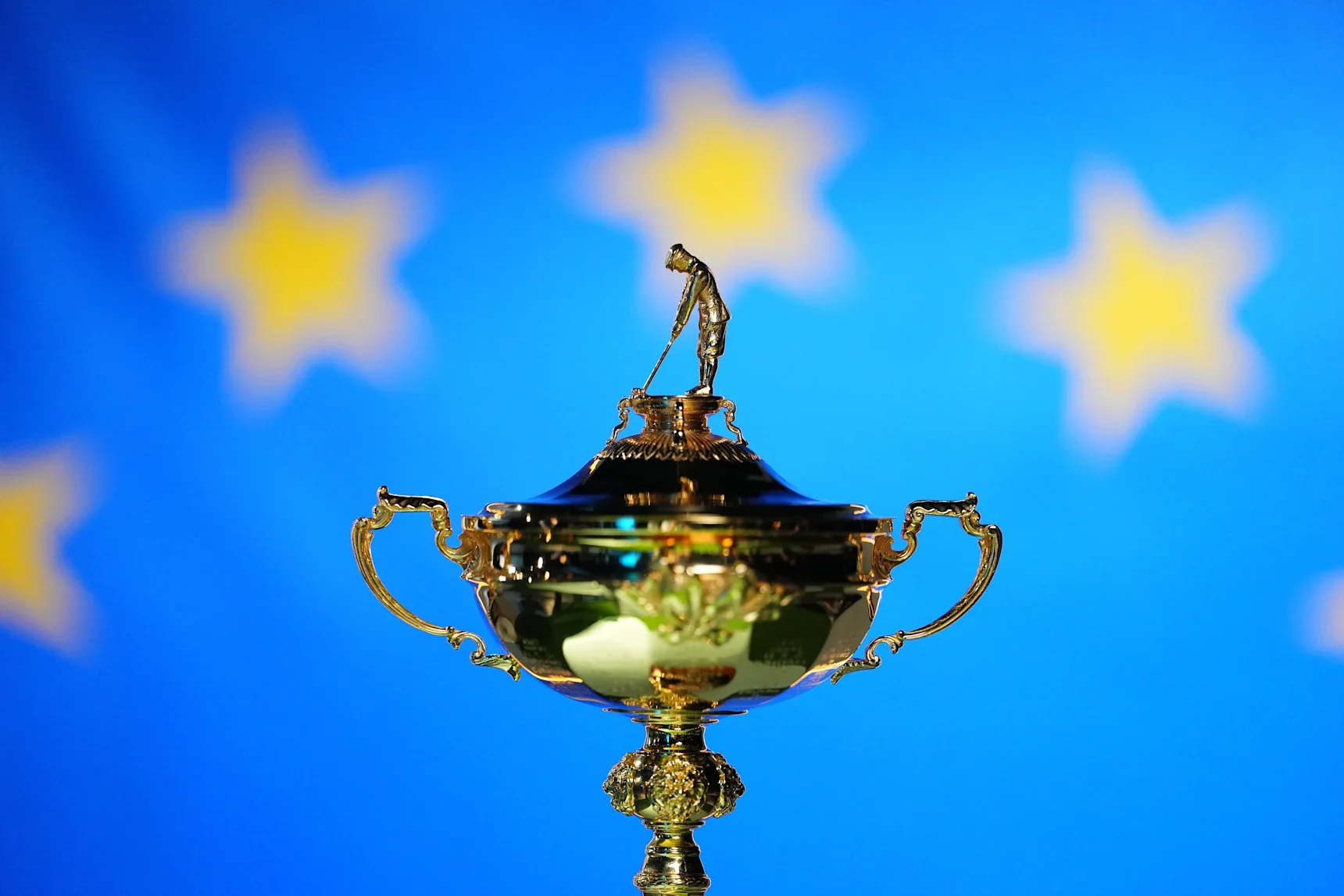 Ryder Cup trophy, a gold cup topped with a golfer figurine, displayed against a European flag backdrop.