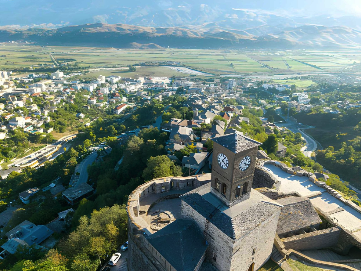 Aerial view of an old stone clock tower in a fortified structure overlooking a small town surrounded by greenery. Scenic mountain landscape in the background. Historic architecture and rural community.