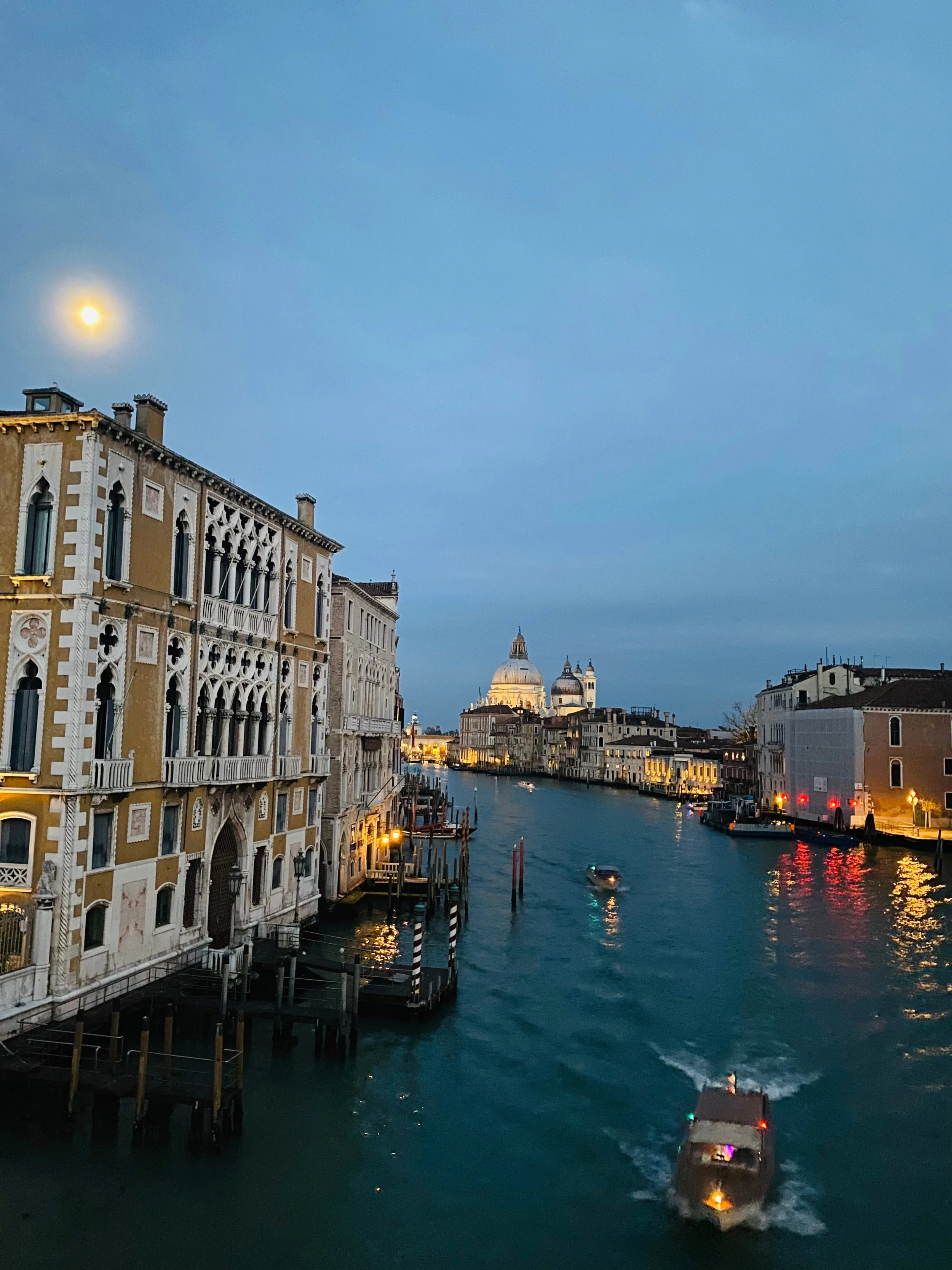 A twilight view of Venice’s Grand Canal from Ponte dell’Accademia, with boats moving along the shimmering water and the illuminated domes of Santa Maria della Salute basilica in the distance.