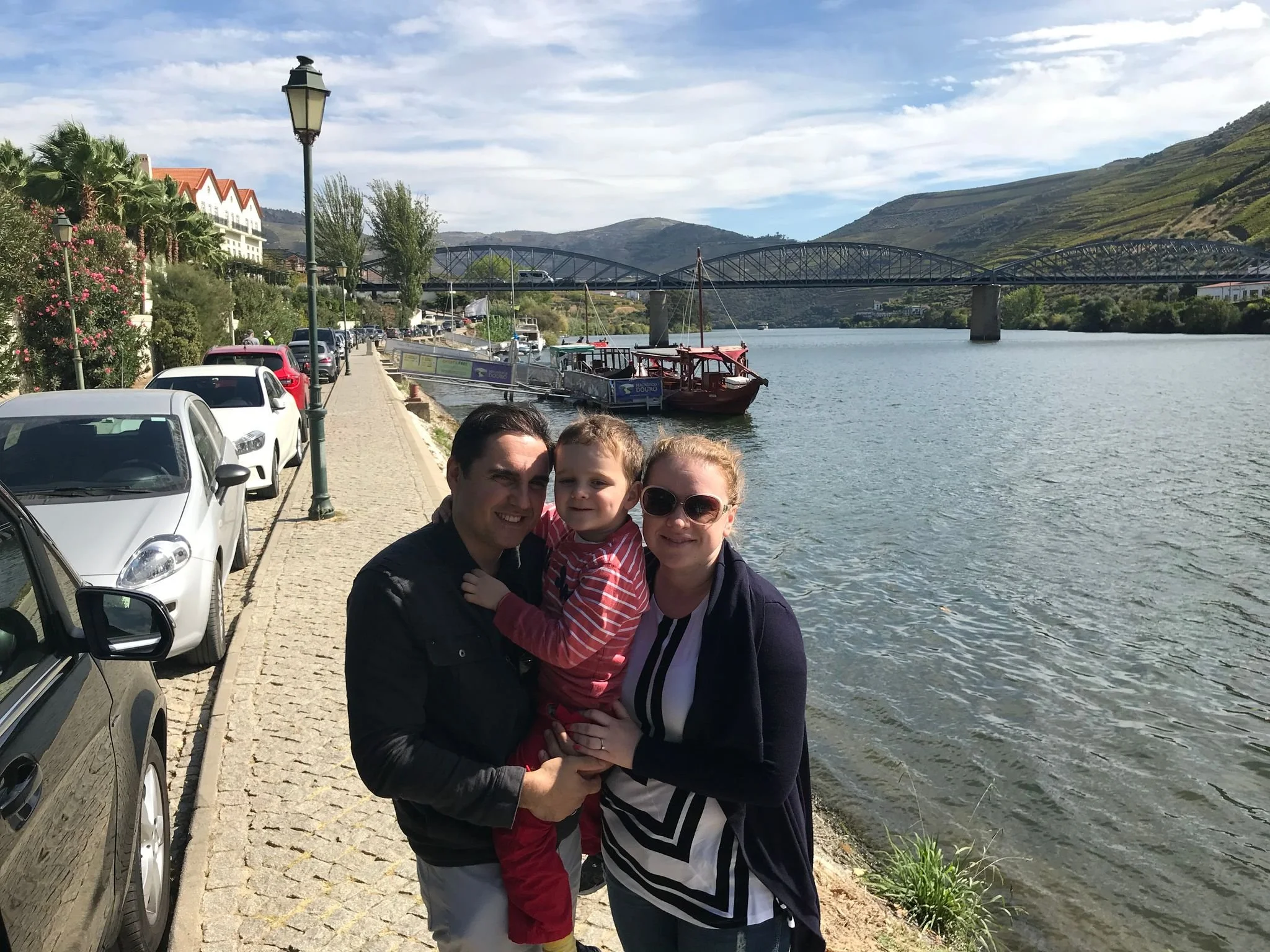 Oleg and Anna family with their first born child posing by the riverfront in Pinhão in the Douro Valley region of Portugal, featuring a scenic bridge, hills, and boats in the background.